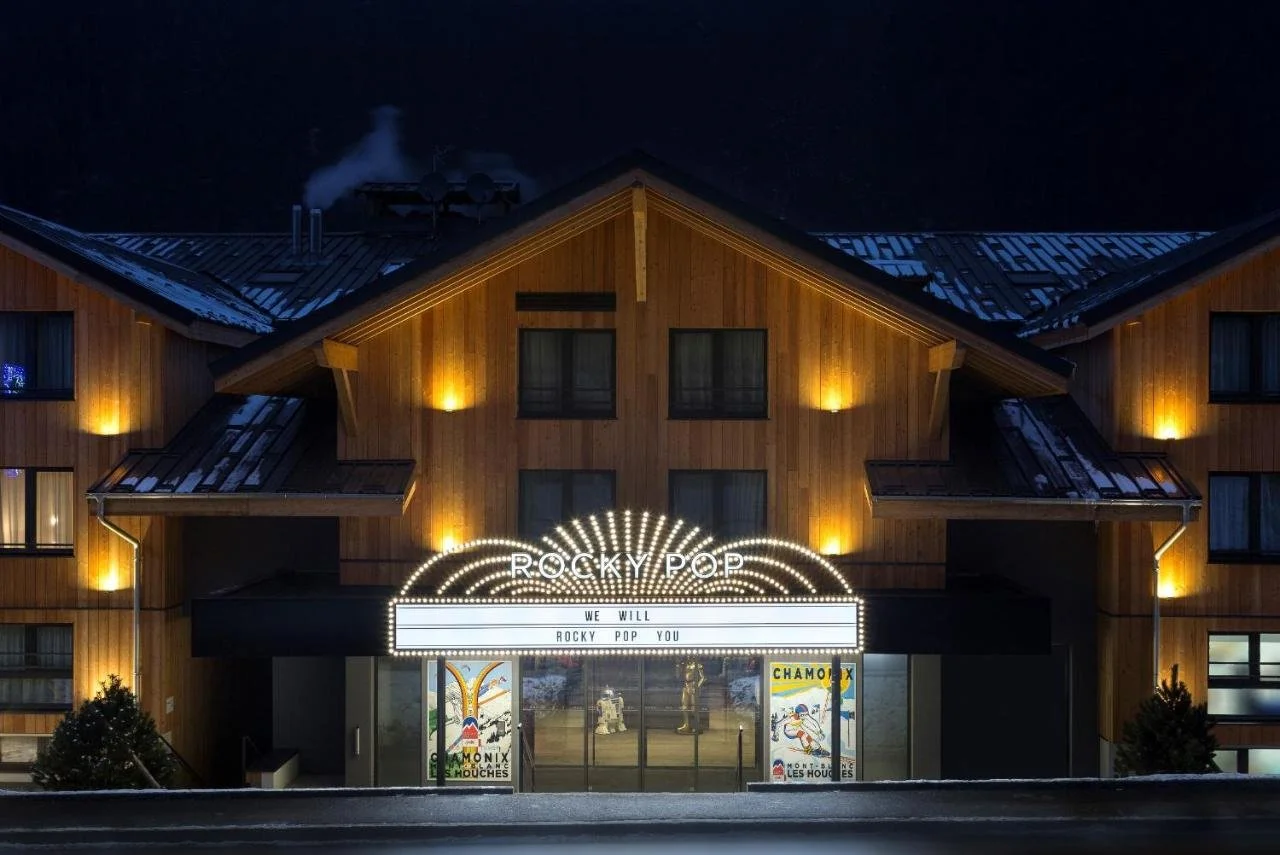 Façade nocturne du Rocky Pop Hotel, avec un panneau de style cinéma et des affiches de ski. Les lumières chaudes du bâtiment en bois et le panneau lumineux créent une ambiance rétro festive.