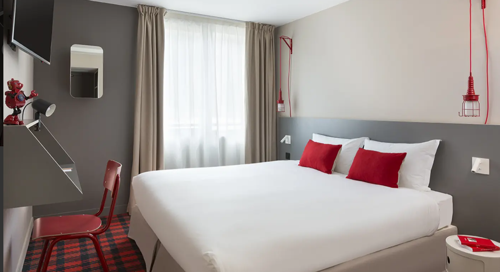 Chambre d’hôtel du Rocky Pop Hotel avec un design moderne, murs gris et accents rouges. Le grand lit avec des oreillers rouges crée une atmosphère accueillante et fonctionnelle.