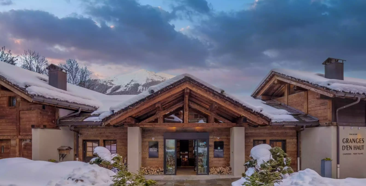 L’entrée de l’Hôtel des Granges d’en Haut aux Houches, luxueux chalet en bois avec neige sur le toit et tas de bois. Les majestueuses montagnes s’élèvent au fond, créant une atmosphère accueillante.