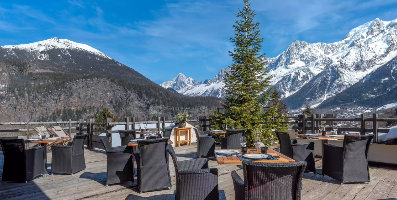 Terrasse extérieure de l’Hôtel Granges d’en Haut avec des tables et des chaises offrant une vue panoramique sur les majestueuses montagnes enneigées des Houches.