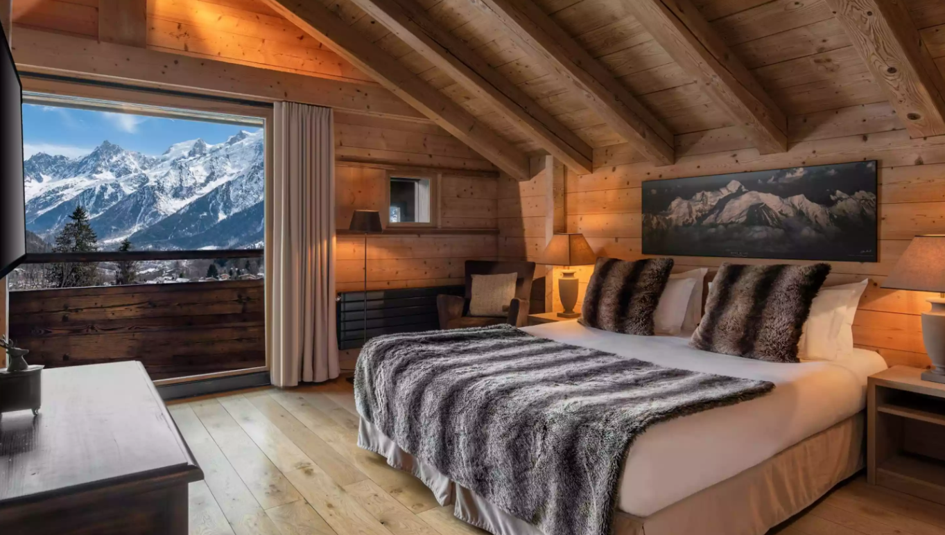 Chambre de luxe à l’Hôtel Granges d’en Haut, avec un design rustique-chic, des murs en bois et un balcon avec vue sur la montagne. L’espace, avec un grand lit et des détails en cuir, est parfait pour le repos.