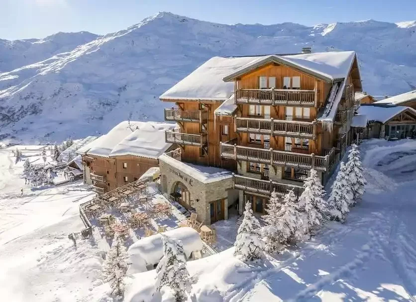 Vue extérieure de l’hôtel Kaya aux Menuires. Le grand chalet en bois et pierre, recouvert de neige, dispose de grands balcons et d’une terrasse extérieure avec tables et chaises.