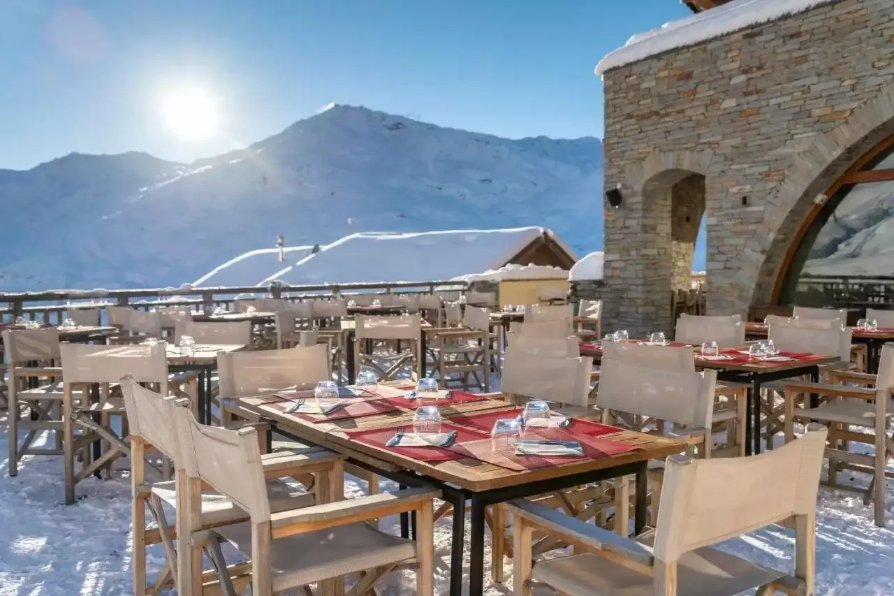 Vue extérieure de l’hôtel Kaya aux Menuires. Le grand chalet en bois et pierre, recouvert de neige, dispose de grands balcons et d’une terrasse extérieure avec tables et chaises.
