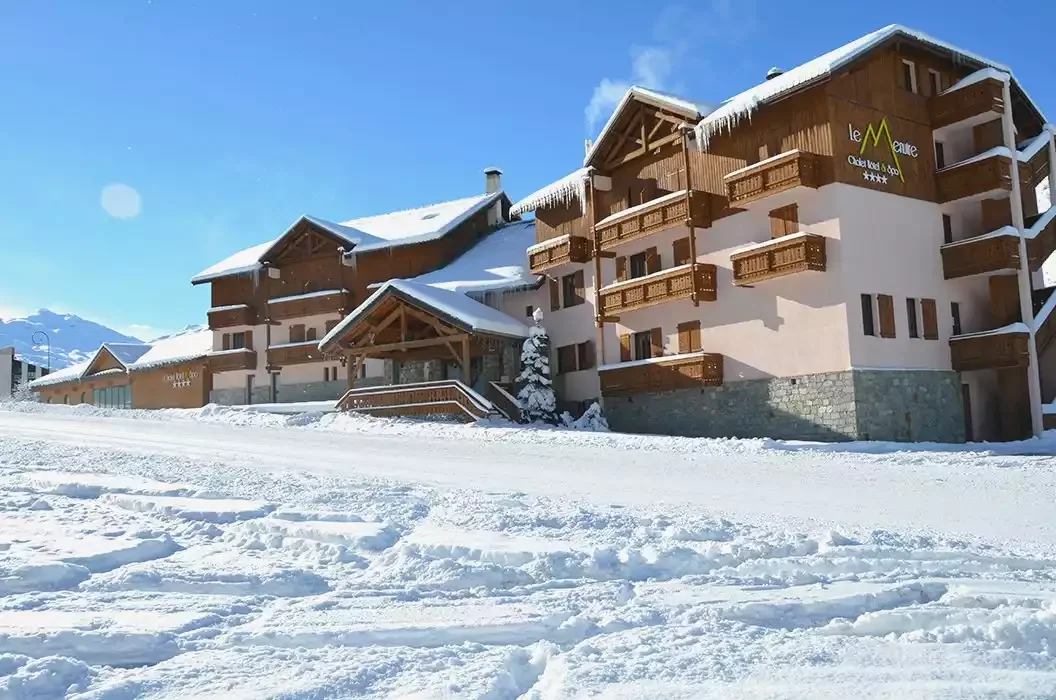 Vue extérieure du **Le Meuire Chalet Hotel & Spa **** par une journée ensoleillée d’hiver. L’hôtel, couvert de neige et de glaçons, a une architecture qui combine la pierre et le bois, avec de grands balcons.