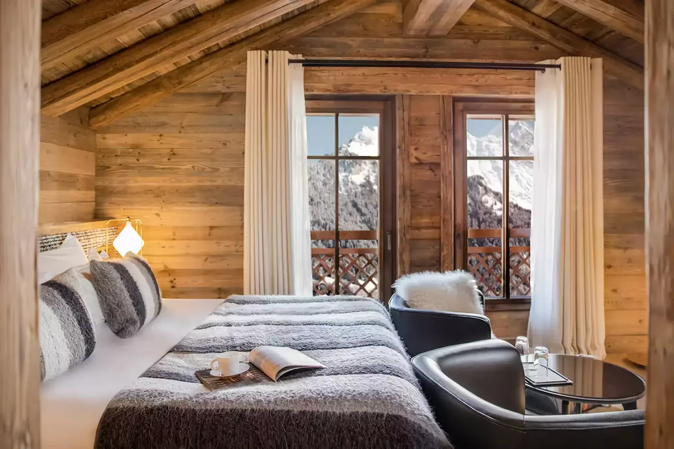 Chambre d’hôtel à Belambra clubs avec un design moderne et confortable. Le lit a une couette à carreaux rouges et une grande fenêtre donnant sur un balcon avec vue sur les montagnes vertes.