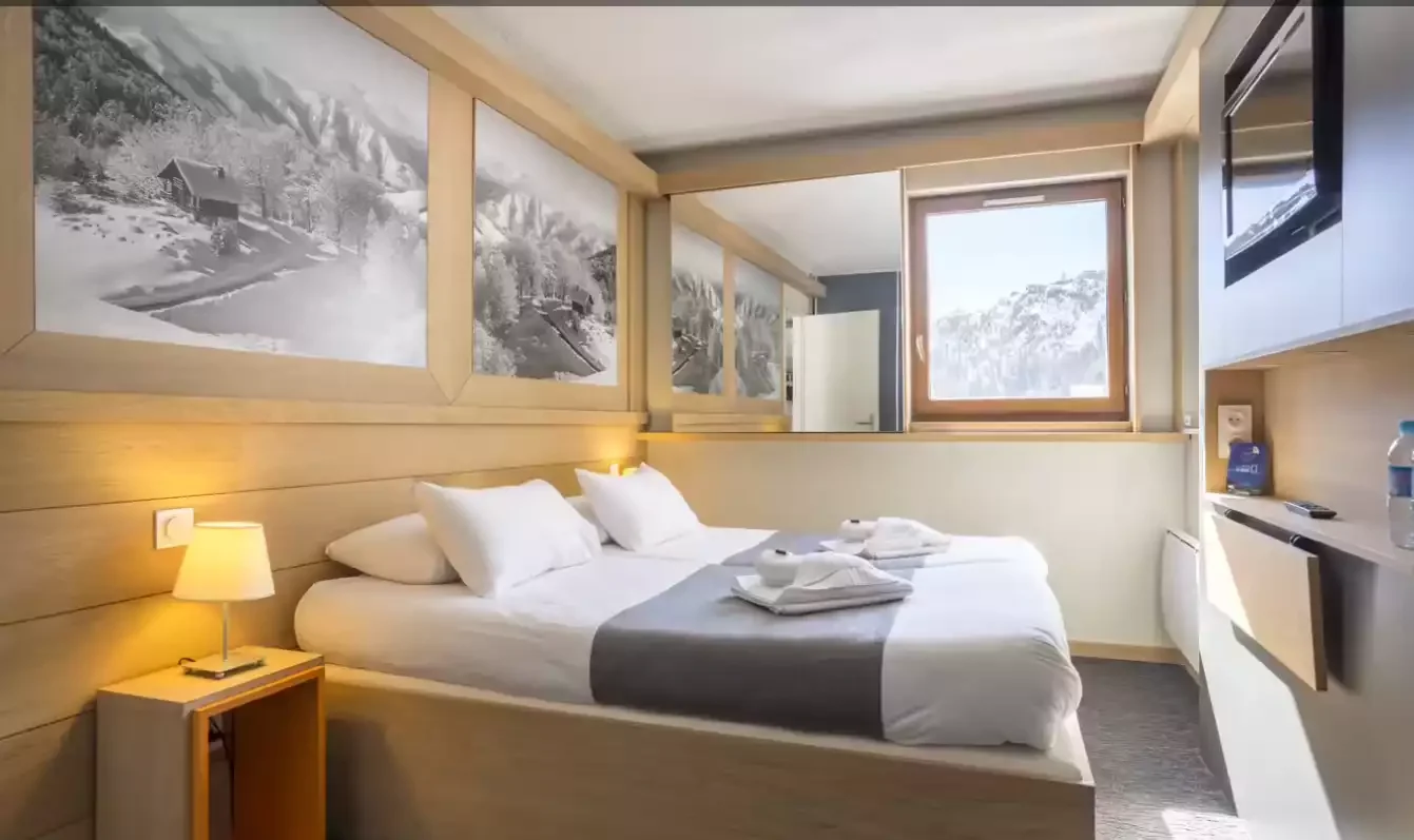 Chambre d’hôtel au design moderne au Club MMV Les Brévières 4*. Le mur en bois et une grande murale de montagne au-dessus du lit créent une atmosphère accueillante.