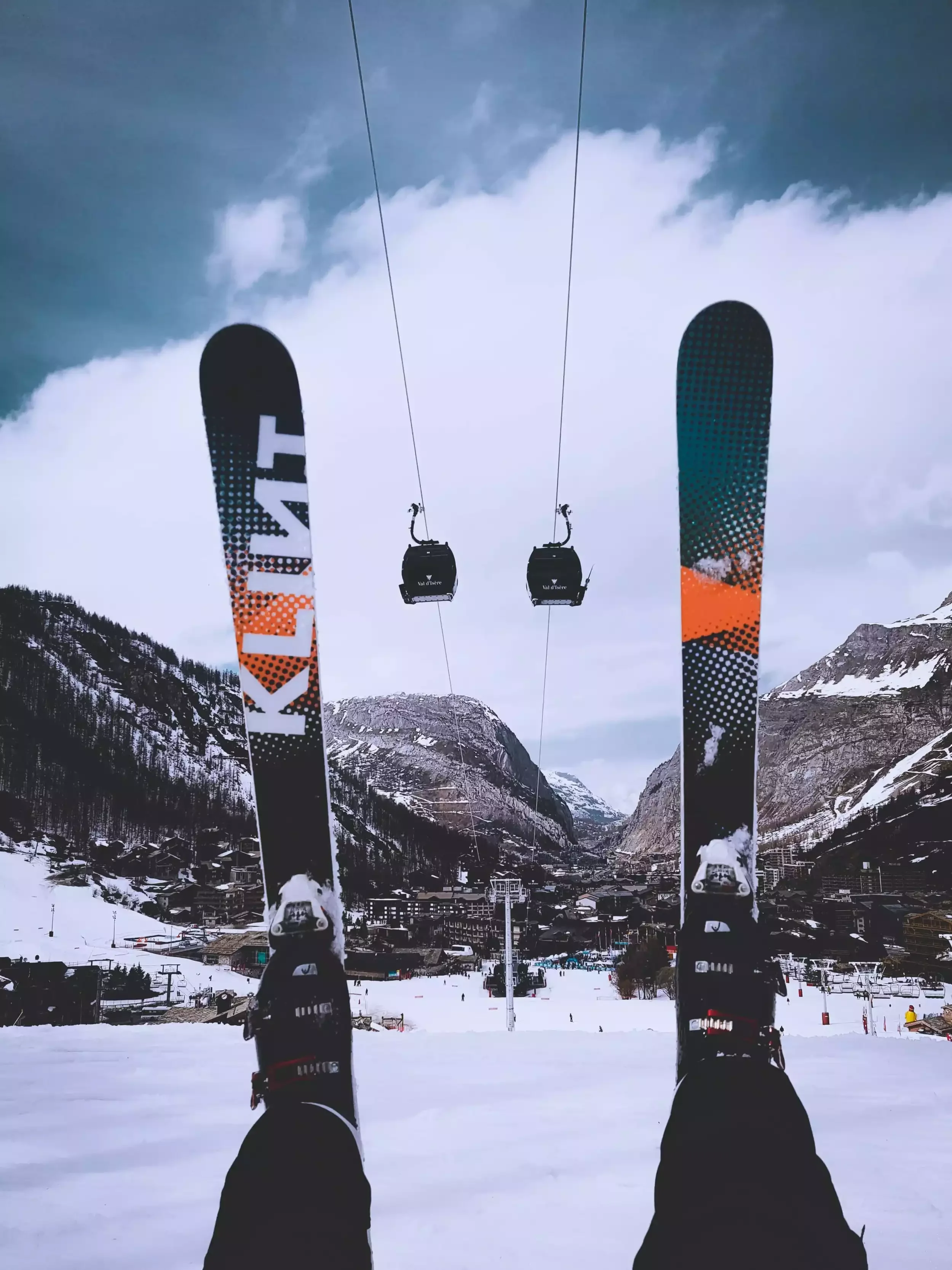 Vue à la première personne d’une station de ski dans le Val d’Isère entre les skis et les bottes d’un skieur. On voit les gondoles et le village enneigé au fond.