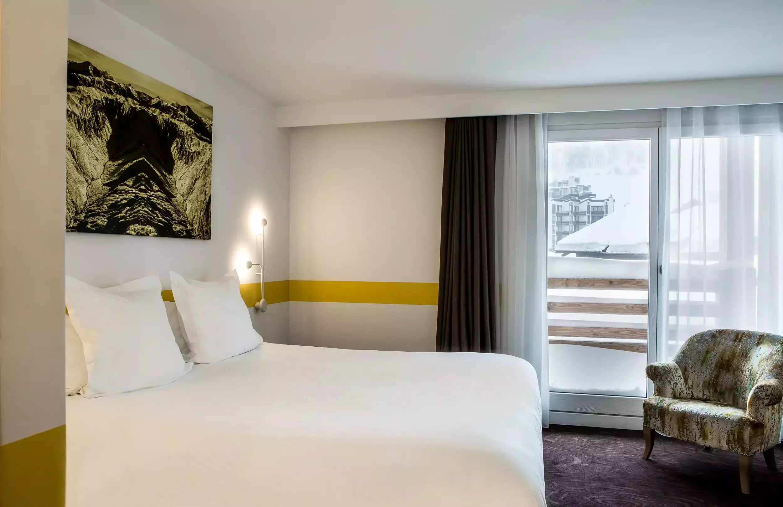 Chambre d’hôtel à l’Hôtel Ormelune avec un grand lit et un design moderne. Une bande jaune sur le mur et une chaise avec un imprimé floral créent un espace accueillant et unique.