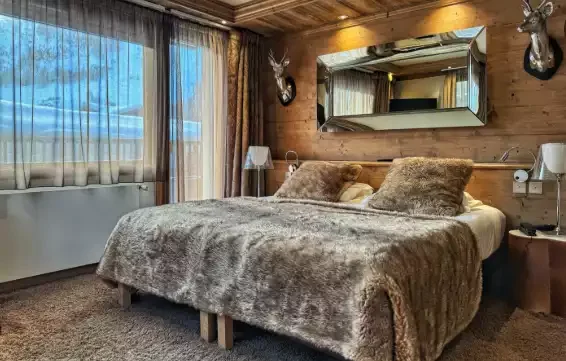 Chambre de luxe à l’Hôtel Tsanteleina avec un design rustique en bois et un grand lit avec une couverture en cuir. La fenêtre donne sur les montagnes, créant une atmosphère chaleureuse et authentique.