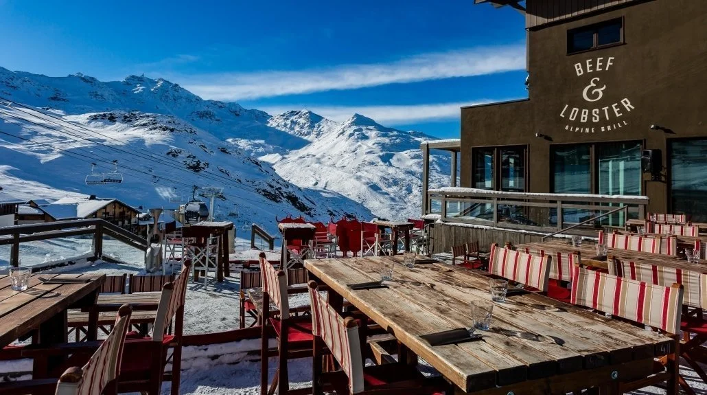 Terrasse extérieure d’un restaurant au Hameau du Kashmir, avec des tables en bois et des chaises rouges sur la neige. C’est un endroit avec des vues spectaculaires sur la montagne, parfait pour un repas ou une pause au soleil.