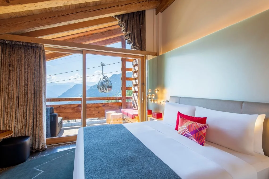 Chambre au design moderne dans le W Verbier hôtel, avec un lit king-size et une grande fenêtre qui offre une vue directe sur une télécabine de ski à Verbier. Une ambiance inspirante pour un séminaire Aylo Montagne.