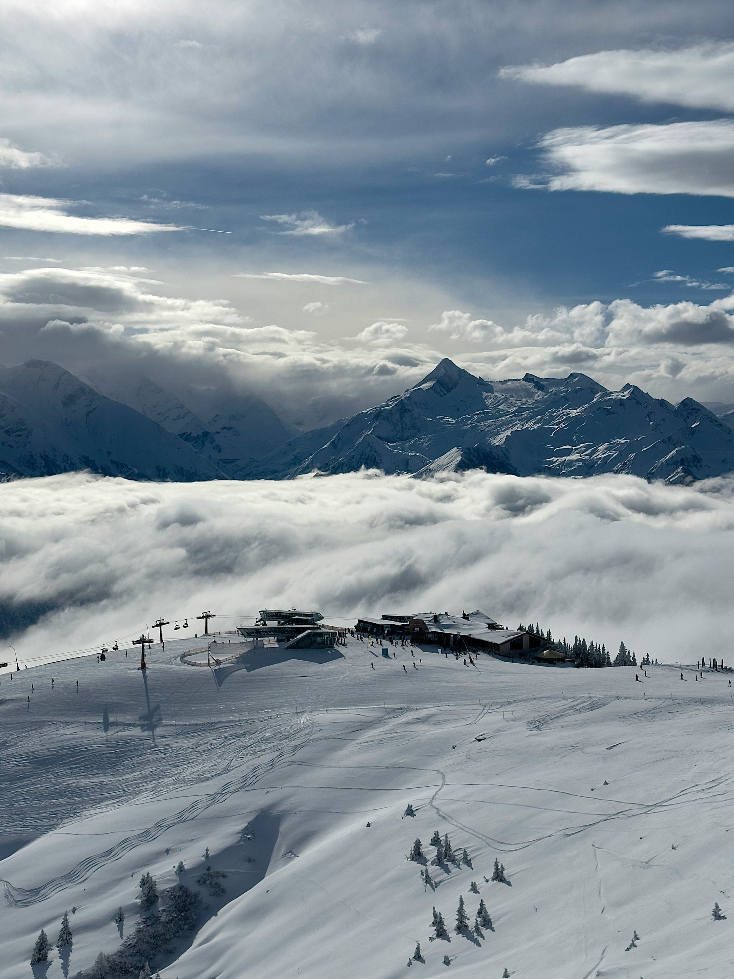 Station de ski en montagne enneigée avec des remontées mécaniques et des skieurs, avec des montagnes enneigées en arrière-plan sous un ciel nuageux.