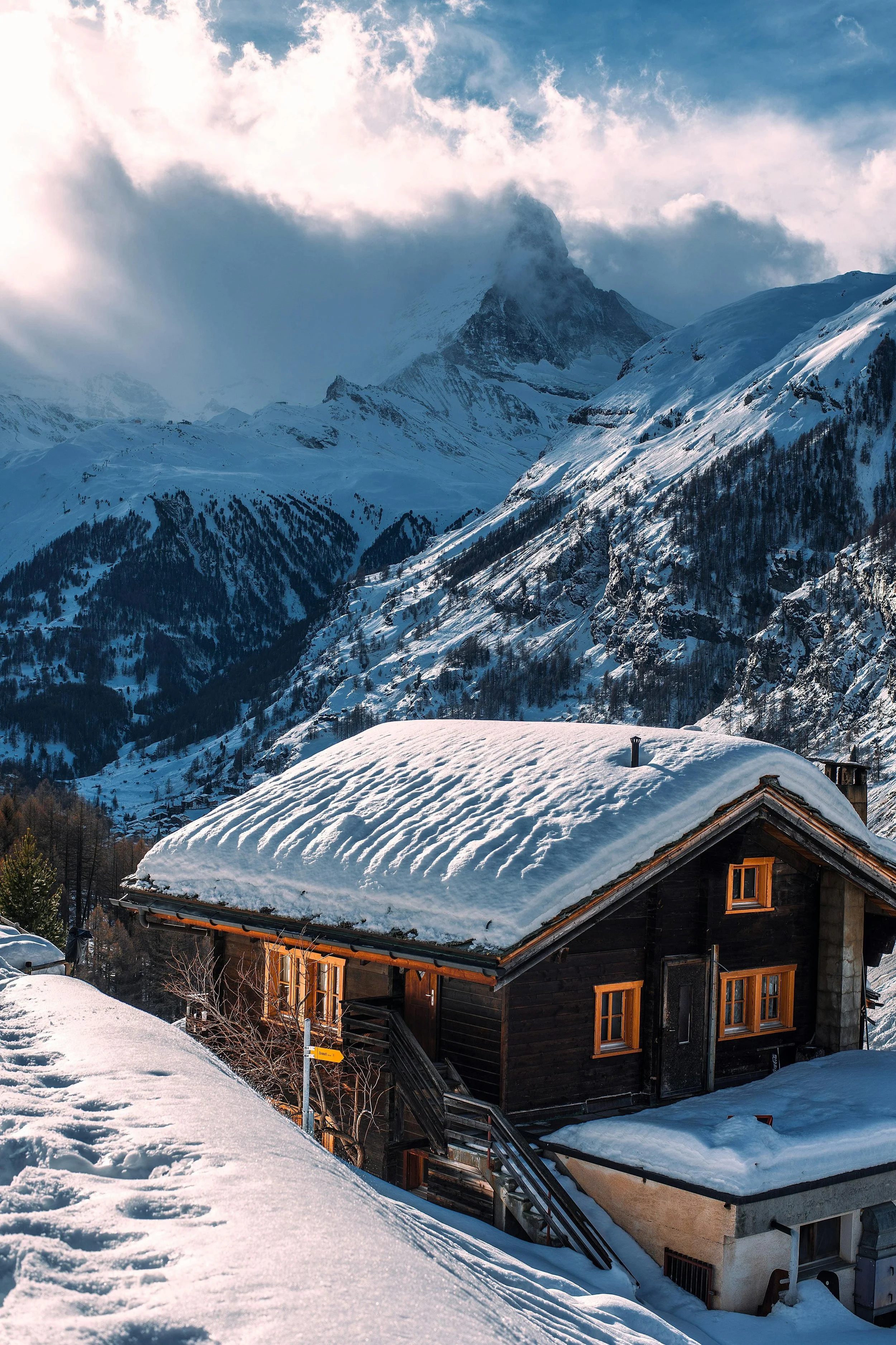 Chalet traditionnel en bois à Zermatt sous la neige, avec les montagnes suisses et le Cervin enveloppé de nuages en arrière-plan.