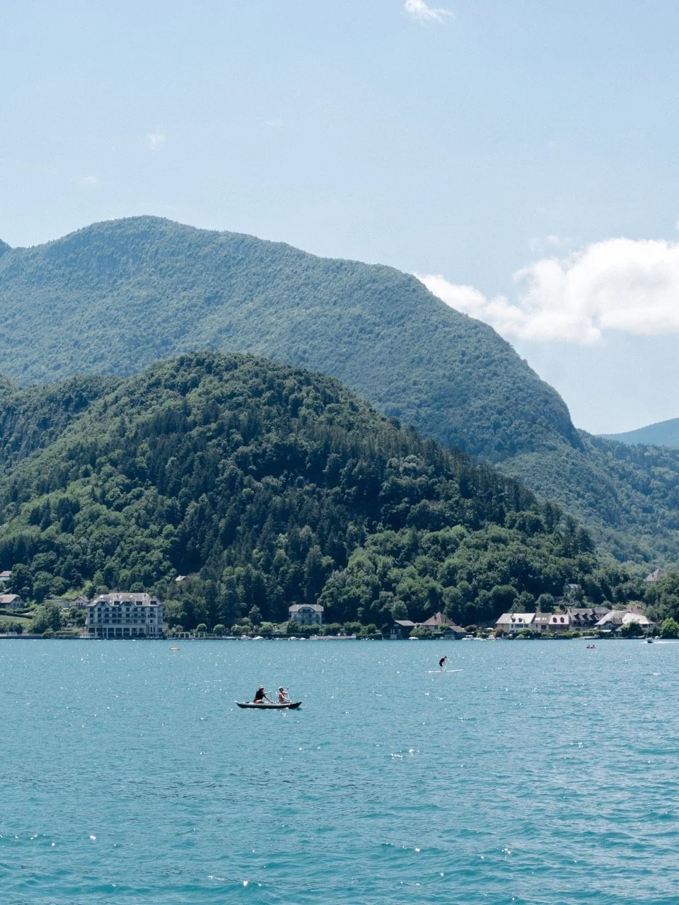 Un lac avec deux personnes dans une petite bateau, montagne verte en arrière-plan et un ciel bleu avec quelques nuages.