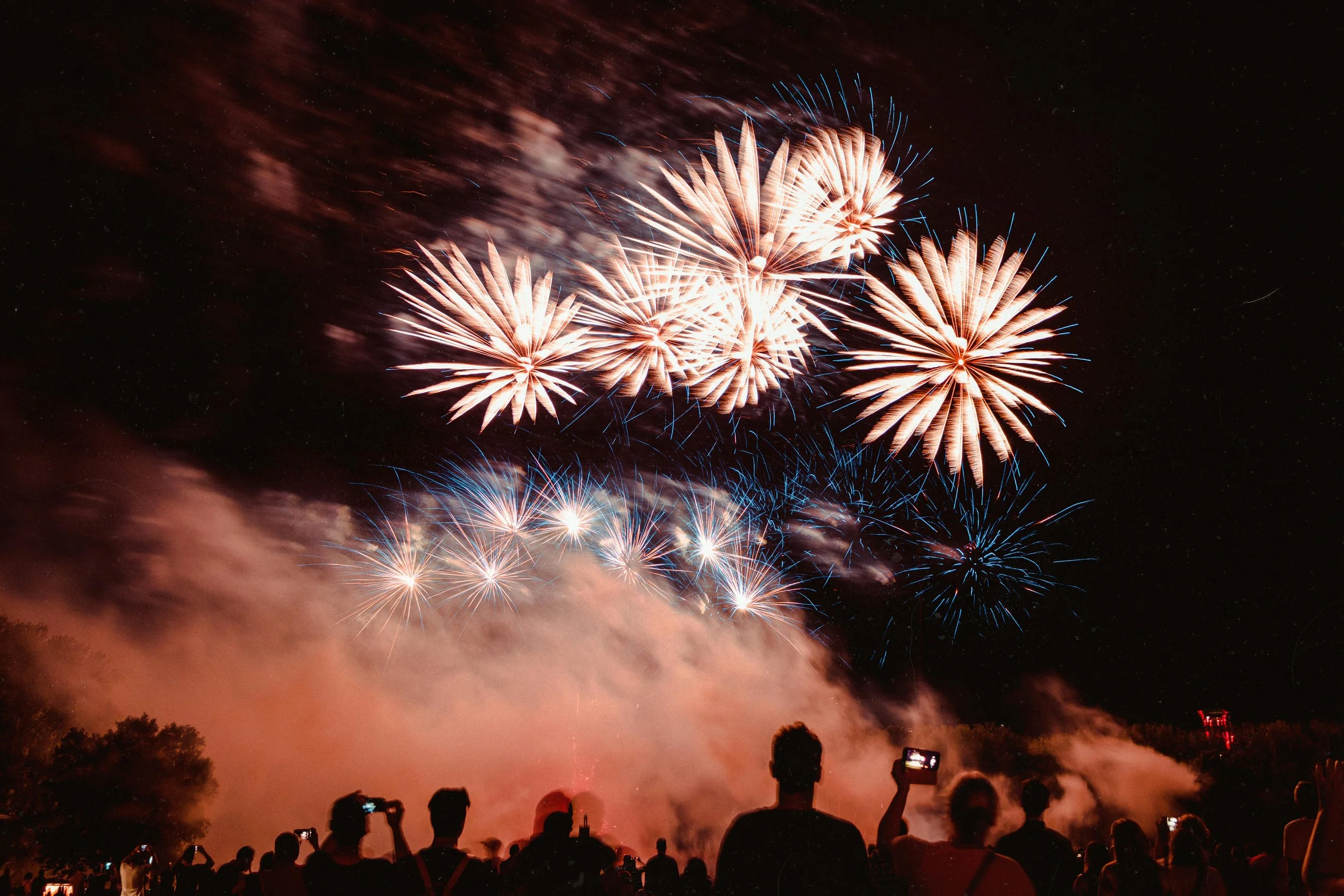Feux d'artifice colorés illuminant le ciel nocturne, avec des spectateurs en silhouette qui prennent des photos.