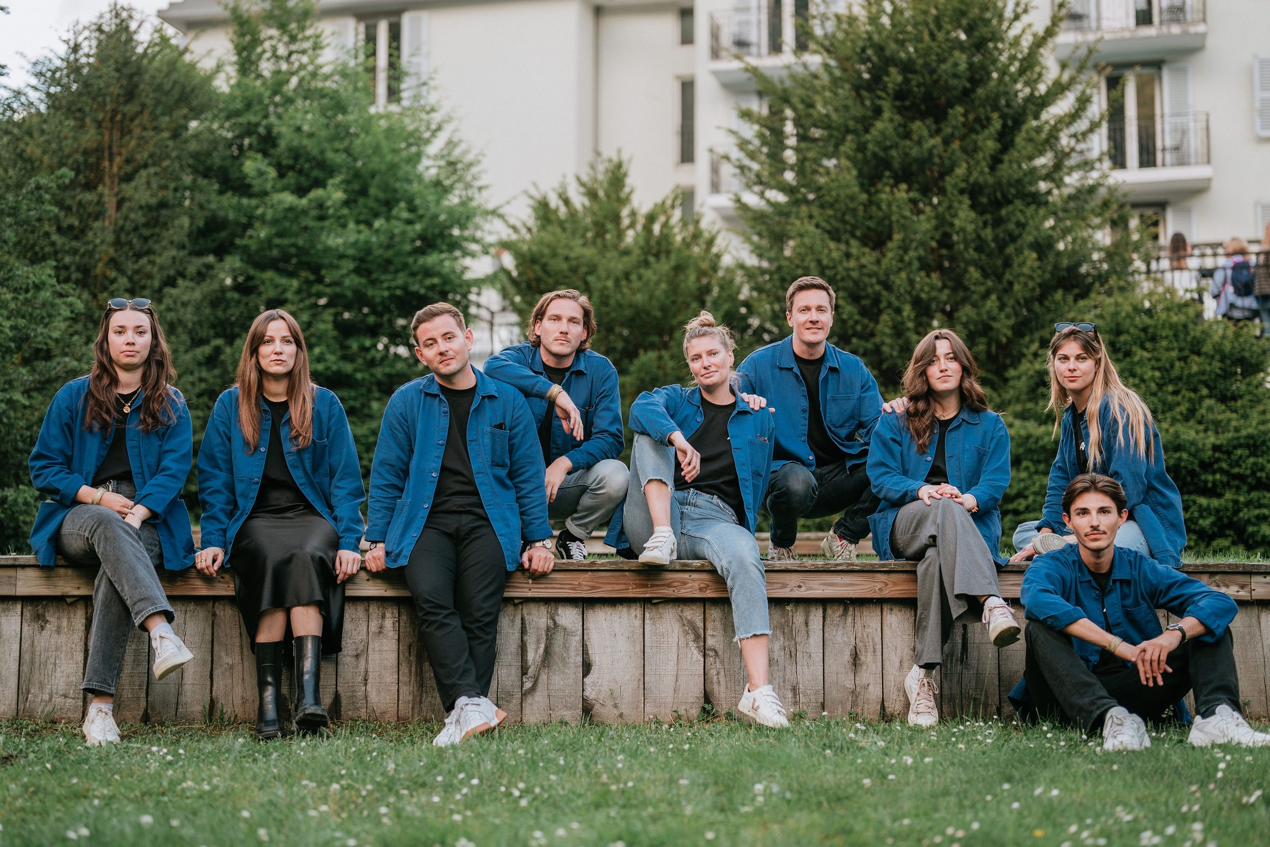Groupe de personnes assises et debout sur une plateforme en bois dans un parc vert, portant des vestes bleues, avec des bâtiments en arrière-plan.