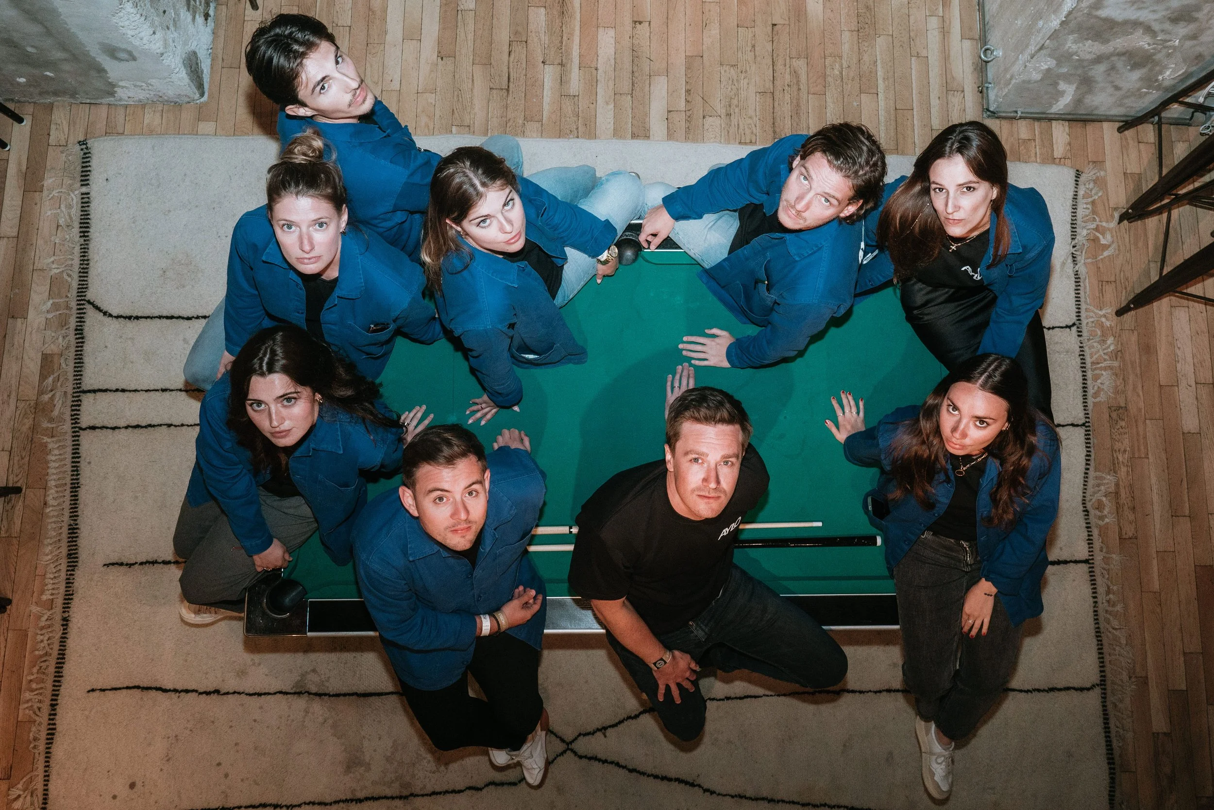 Groupe de neuf jeunes adultes assemblés autour d'une table de ping-pong, vus de dessus, dans une pièce au sol en parquet et tapis beige, portant des vêtements décontractés et regardant vers le haut.