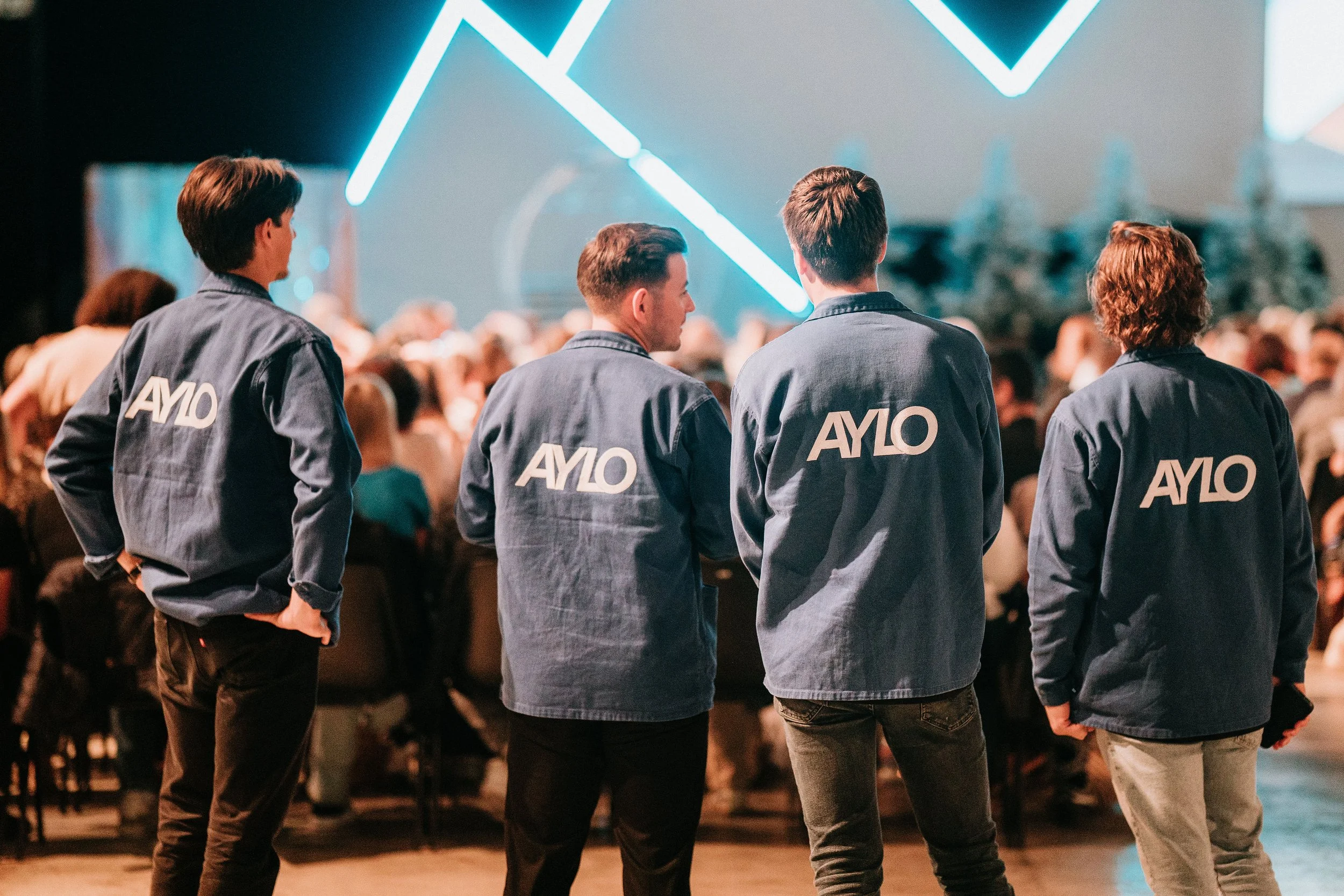 Quatre jeunes hommes portant des vestes avec le logo 'AYLO' observant un événement devant un grand écran avec un fond lumineux futuriste, dans une salle pleine de spectateurs.