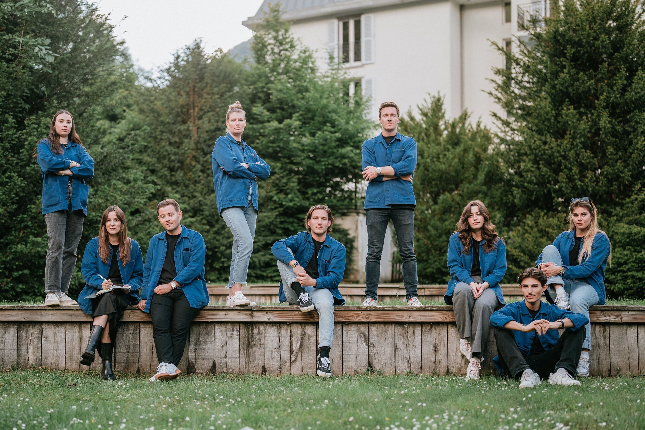 Groupe de jeunes adultes posant en extérieur, certains debout sur une plateforme en bois, d'autres assis devant, avec des arbres et un bâtiment en arrière-plan.