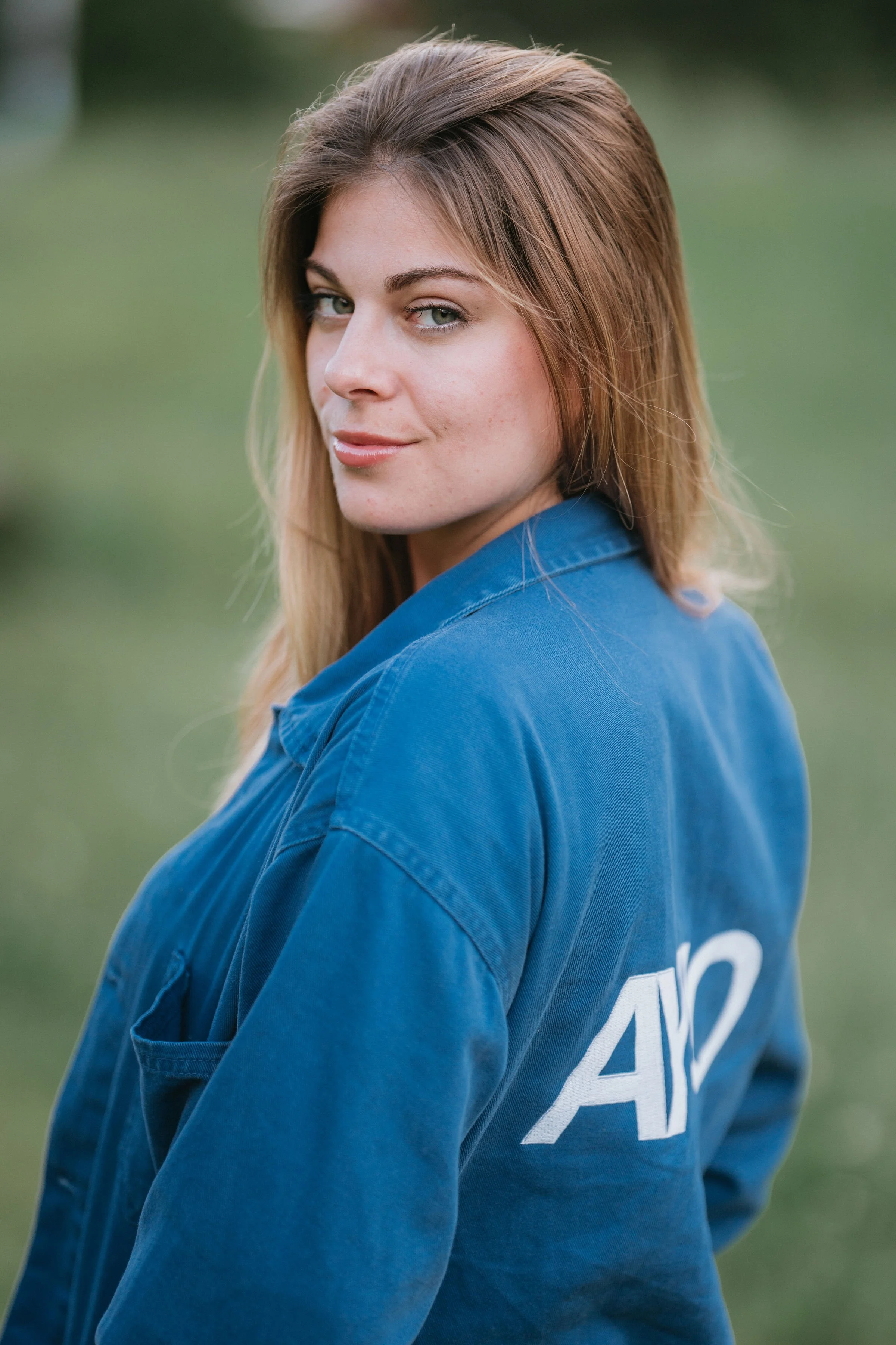 Une jeune femme aux cheveux bruns, yeux verts, portant une veste bleue, regardant par-dessus son épaule, arrière-plan flou vert.