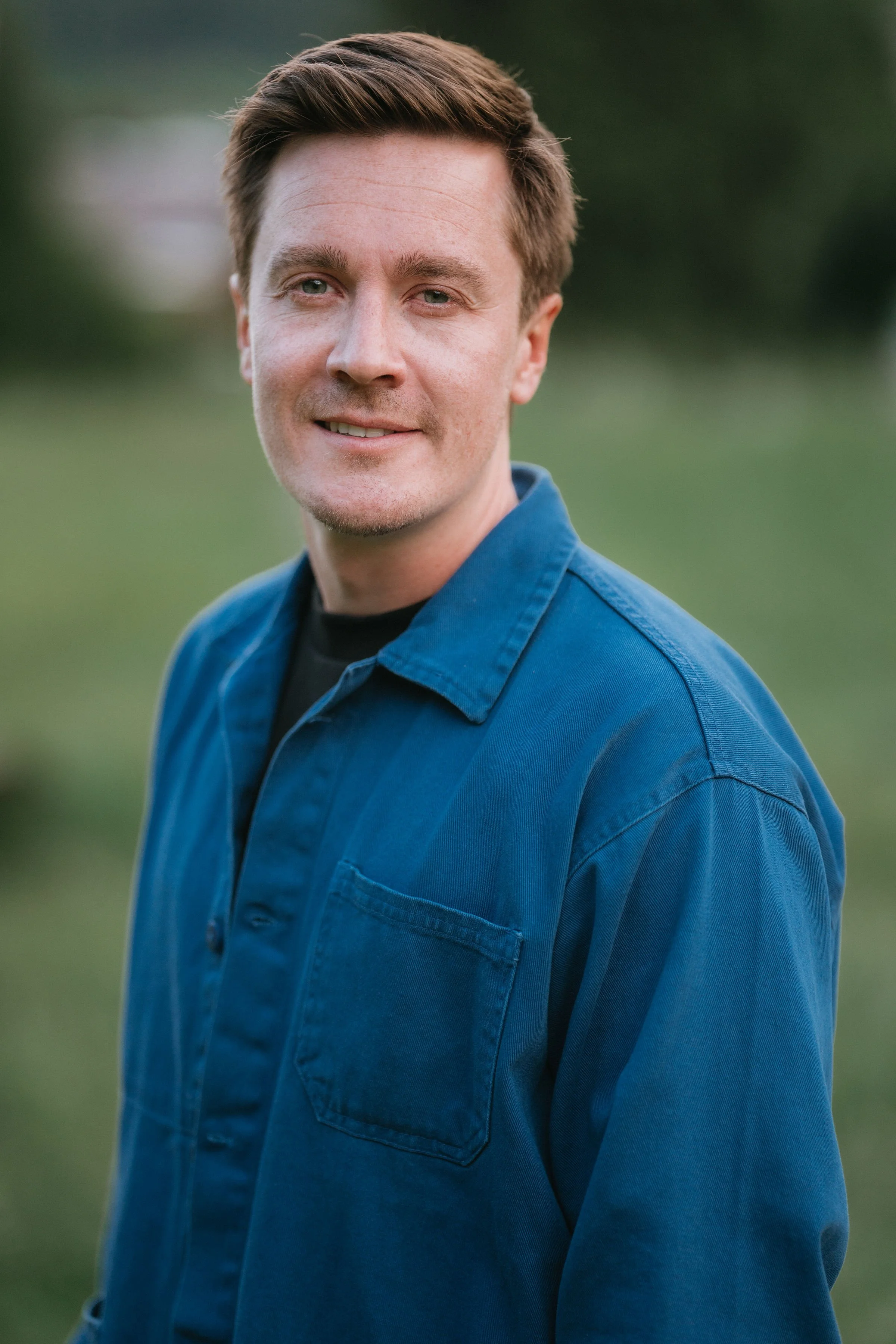 Un homme portant une chemise bleue, souriant légèrement, en extérieur avec un arrière-plan flou de nature.