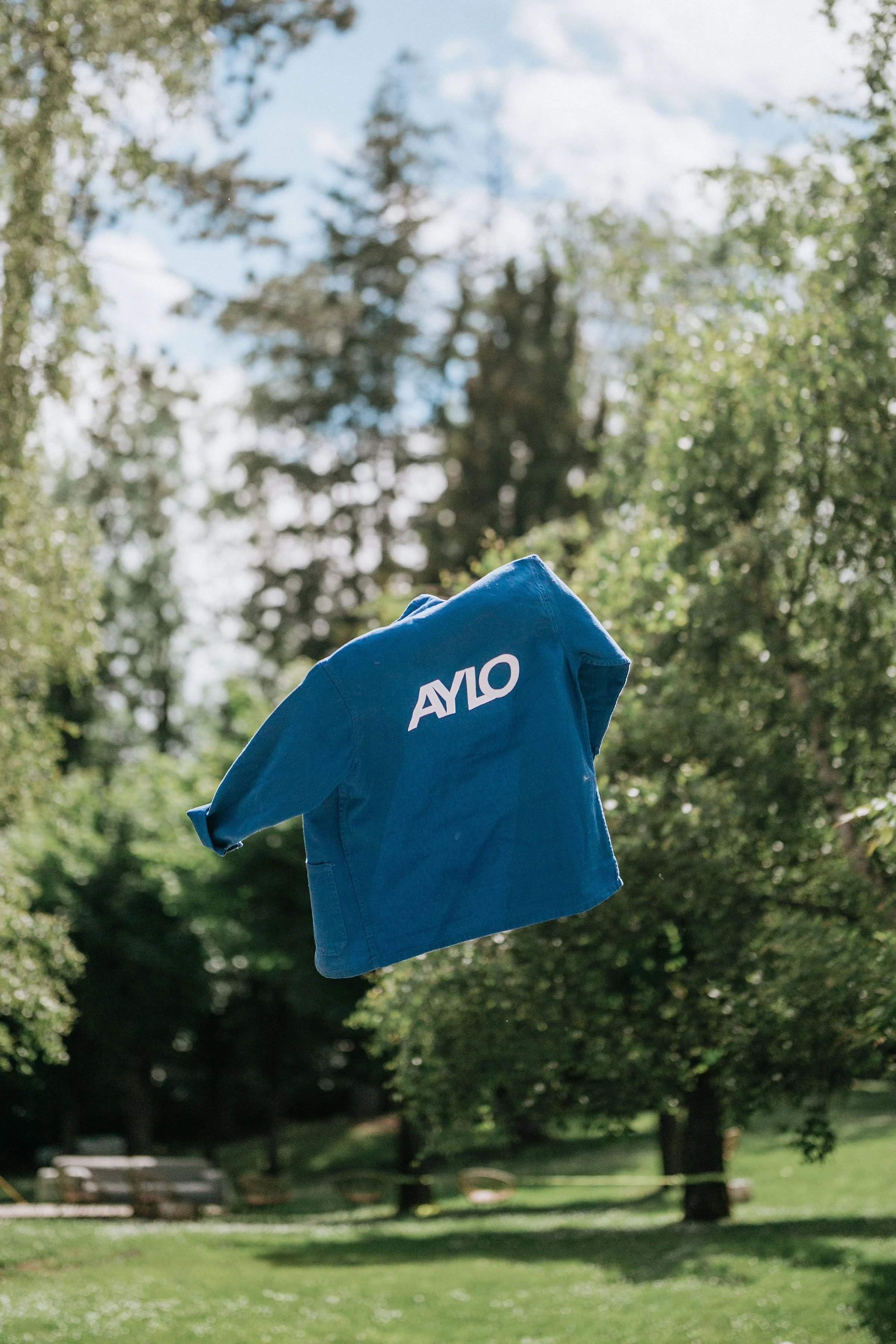 Veste bleue avec le logo 'AYLO' flottant dans les airs dans un parc avec des arbres et un ciel en arrière-plan.