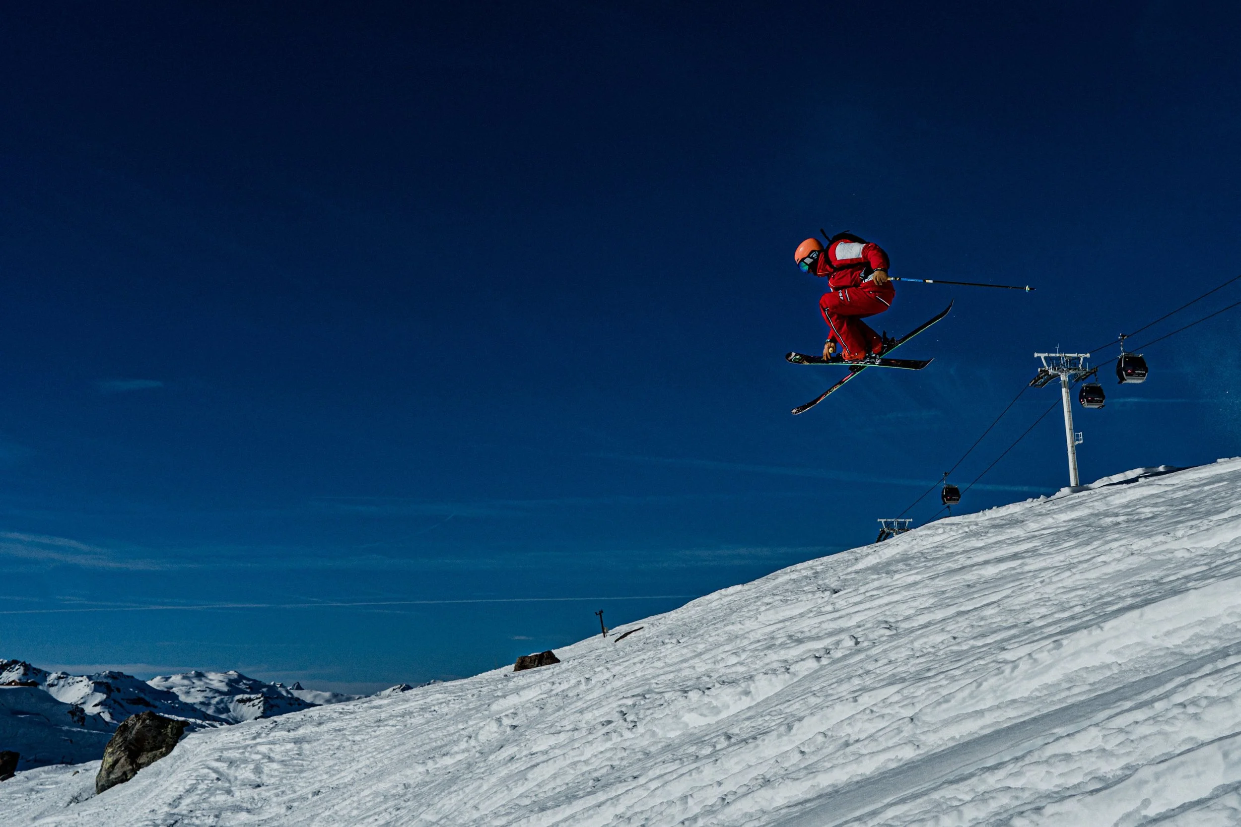 Un skieur en tenue rouge effectue un saut dans la neige, avec un ciel étoilé en arrière-plan et des montagnes enneigées au loin, à côté d'une télécabine.