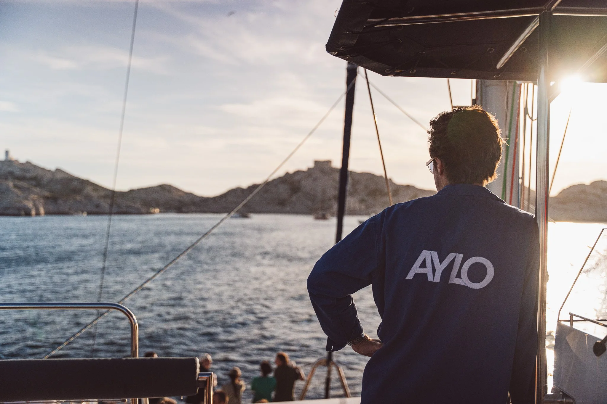 Personne vue de dos sur un bateau, en train de regarder la mer avec des montagnes en arrière-plan, pendant un coucher de soleil.