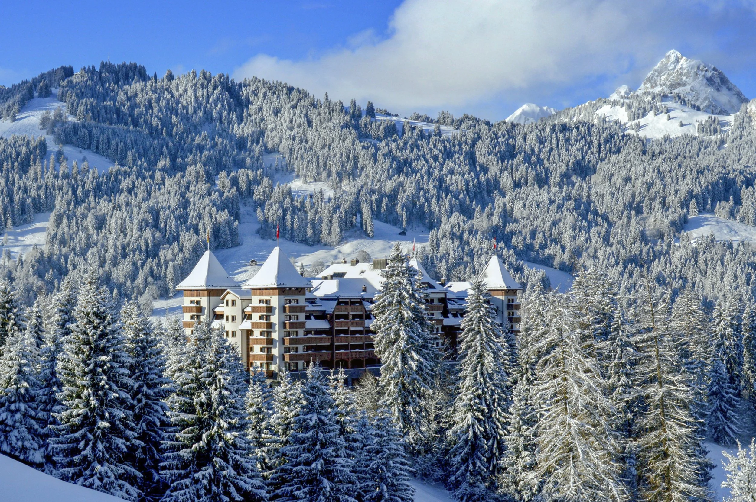 Vue extérieure de l’hôtel The Alpina Gstaad en hiver, entouré de forêts enneigées et des Alpes suisses – Aylo Montagne.