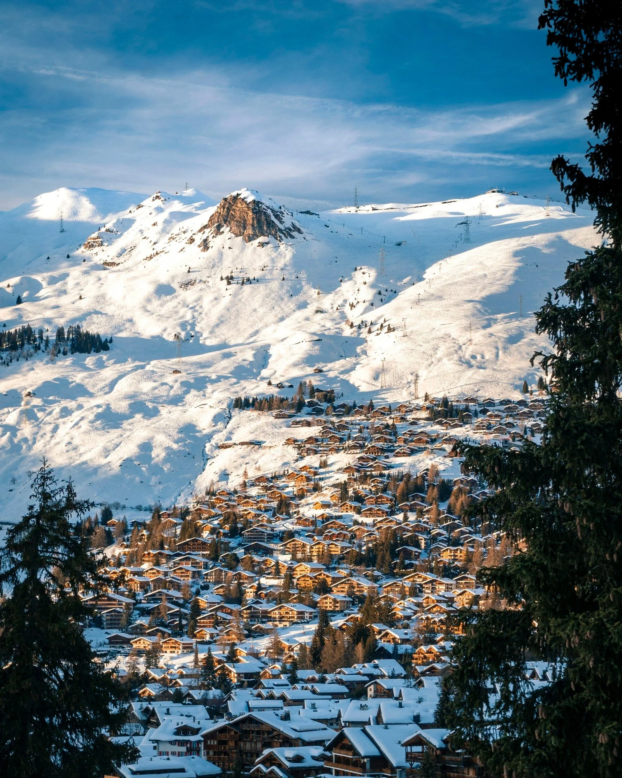 Vue panoramique de la station de ski de Verbier, montrant des centaines de chalets et bâtiments en bois sous la lumière du coucher de soleil. La montagne enneigée s’élève majestueusement au-dessus du village.