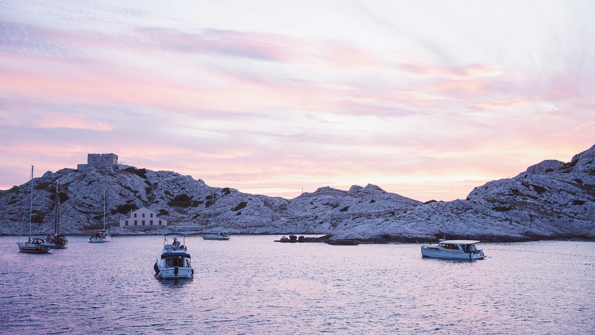 Couchers de soleil pastel sur une baie avec des bateaux amarrés, des rochers blancs et un château en hauteur au sommet d'une colline rocheuse.