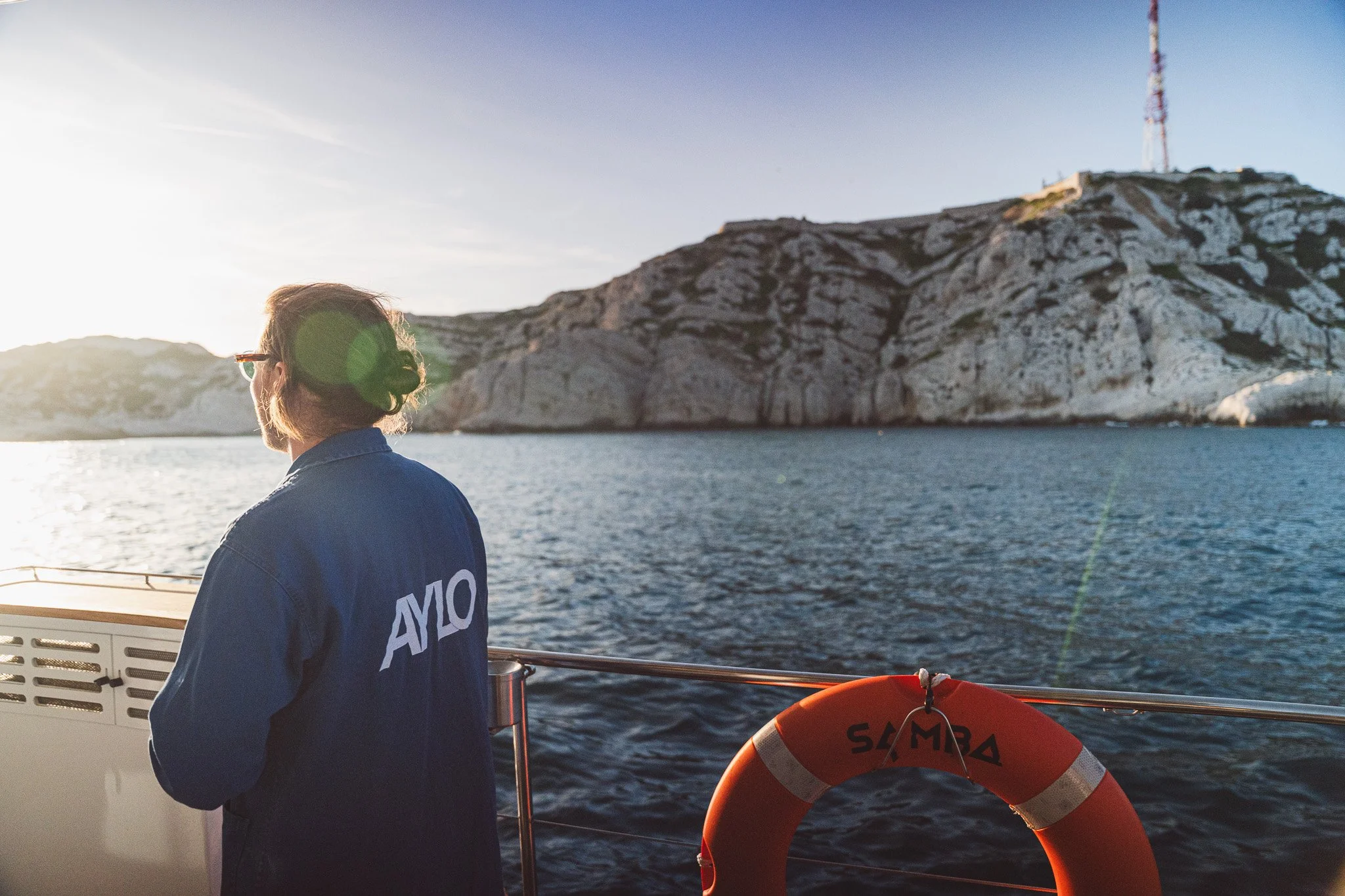 Une personne vue de dos sur un bateau, portant une veste avec le mot 'AMO' écrit dessus, regardant vers une île rocheuse avec une tour de communication au sommet, en mer au coucher du soleil.