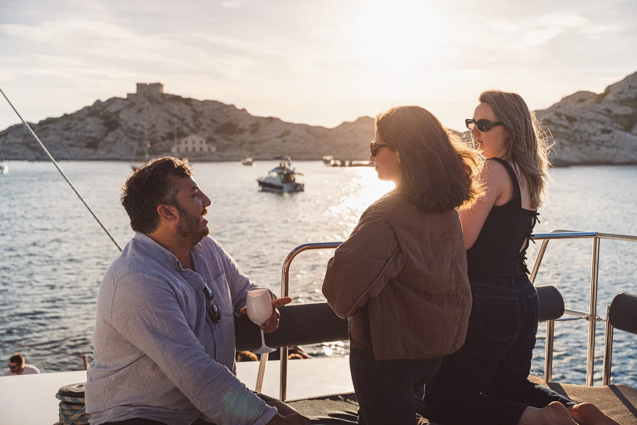 Trois personnes se parlent sur un bateau avec un paysage marin au coucher du soleil en arrière-plan.