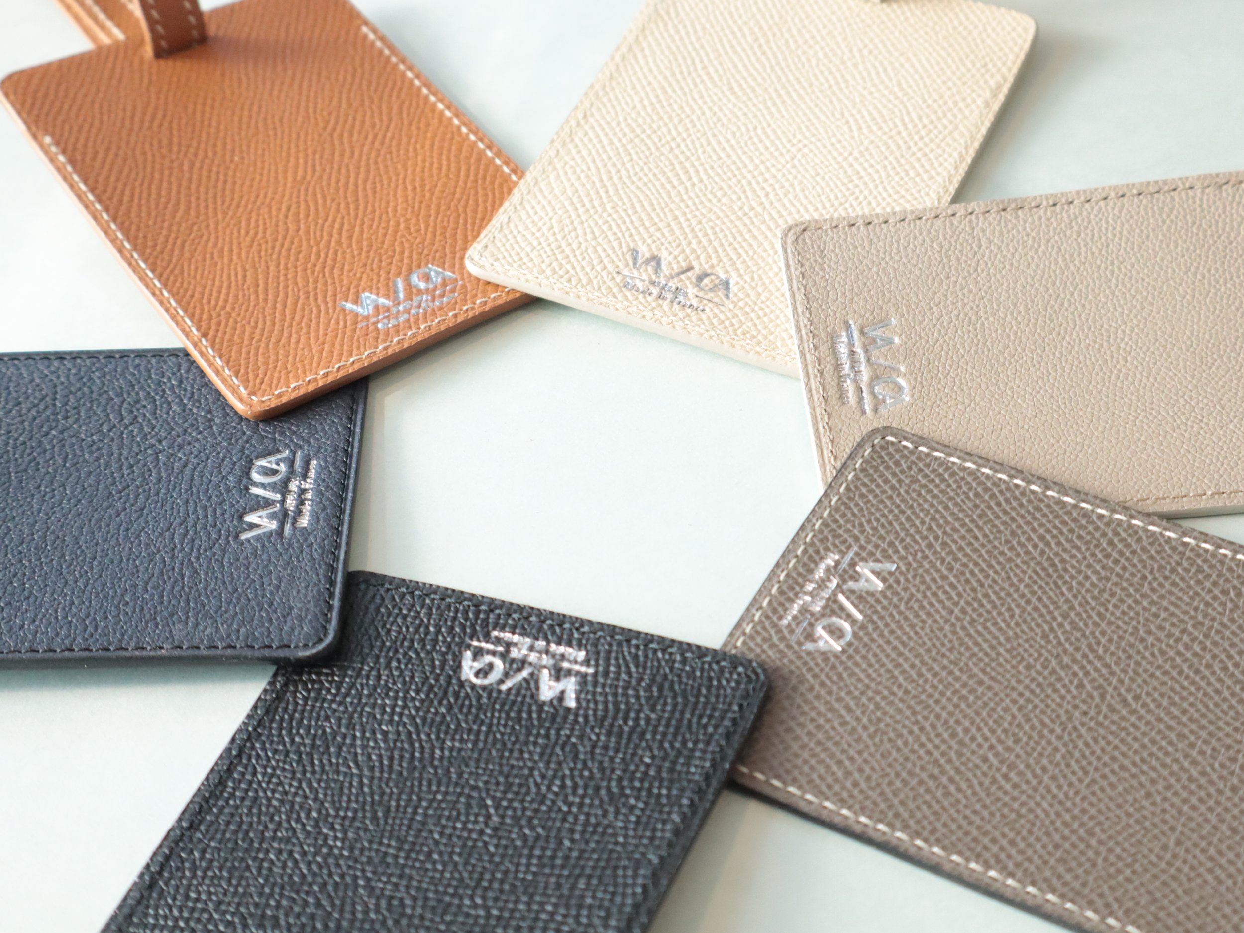 Différents porte-cartes en cuir de plusieurs couleurs disposés en cercle, chacun avec la marque 'VELA' en lettres argentées.