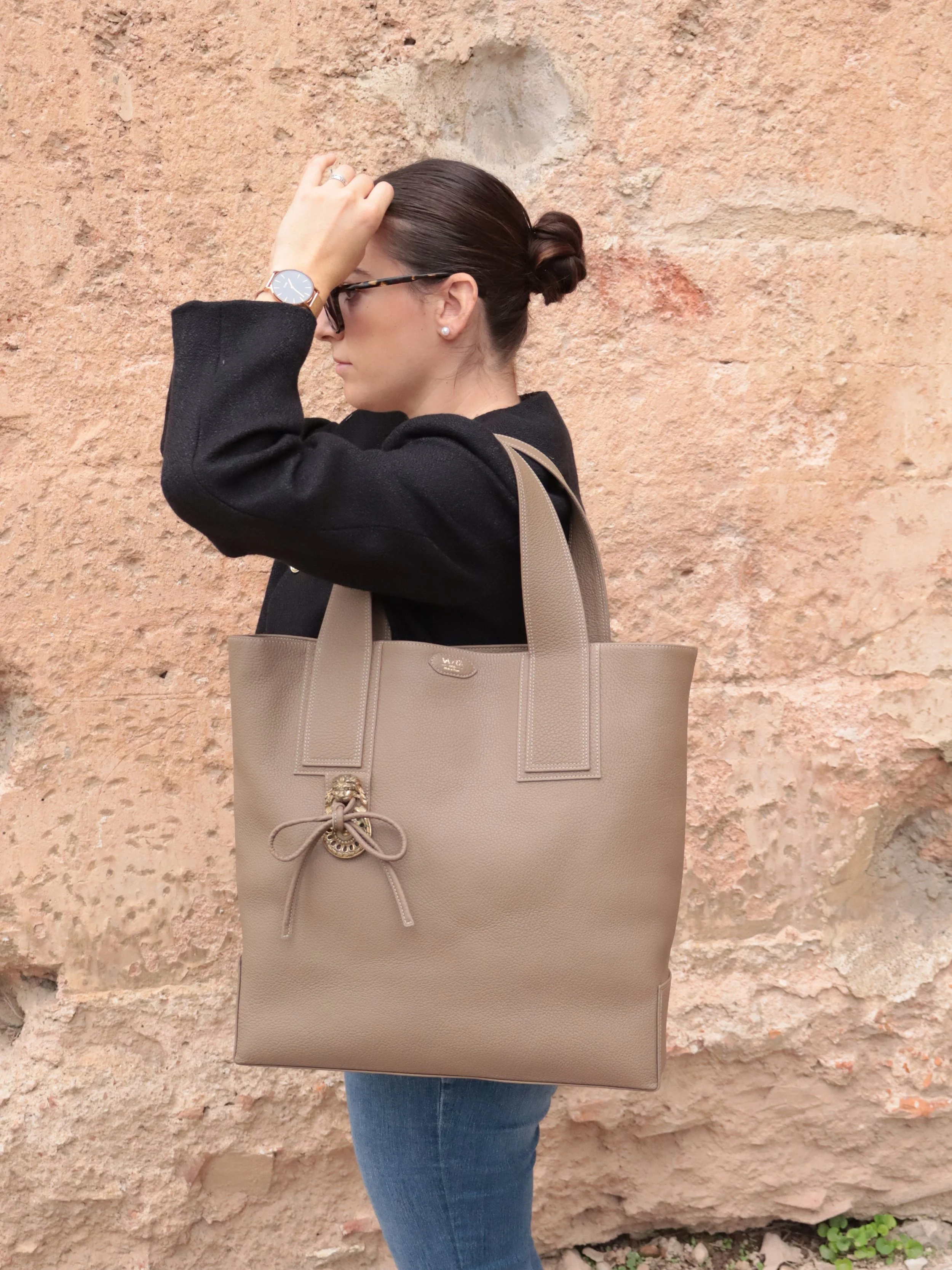 Femme portant des lunettes de soleil, un sac beige en cuir, un jean bleu et un manteau noir, debout contre un mur en pierre couleur terre cuite.