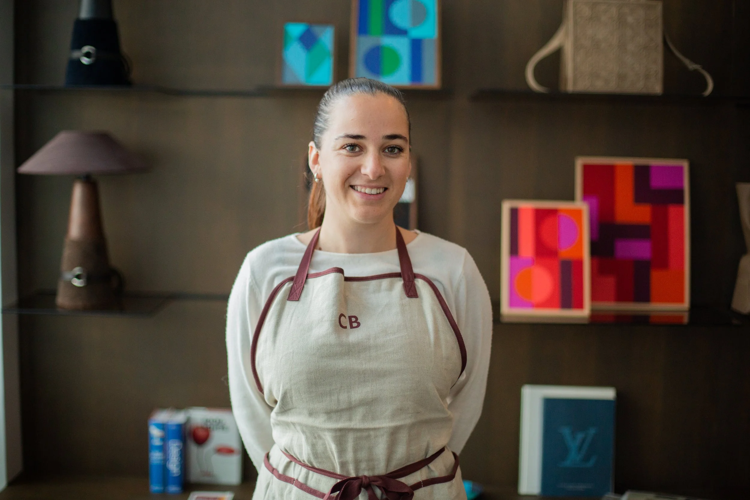 Jeune femme souriante portant un tablier beige avec des détails marron, dans une boutique de décoration avec des tableaux colorés en arrière-plan.