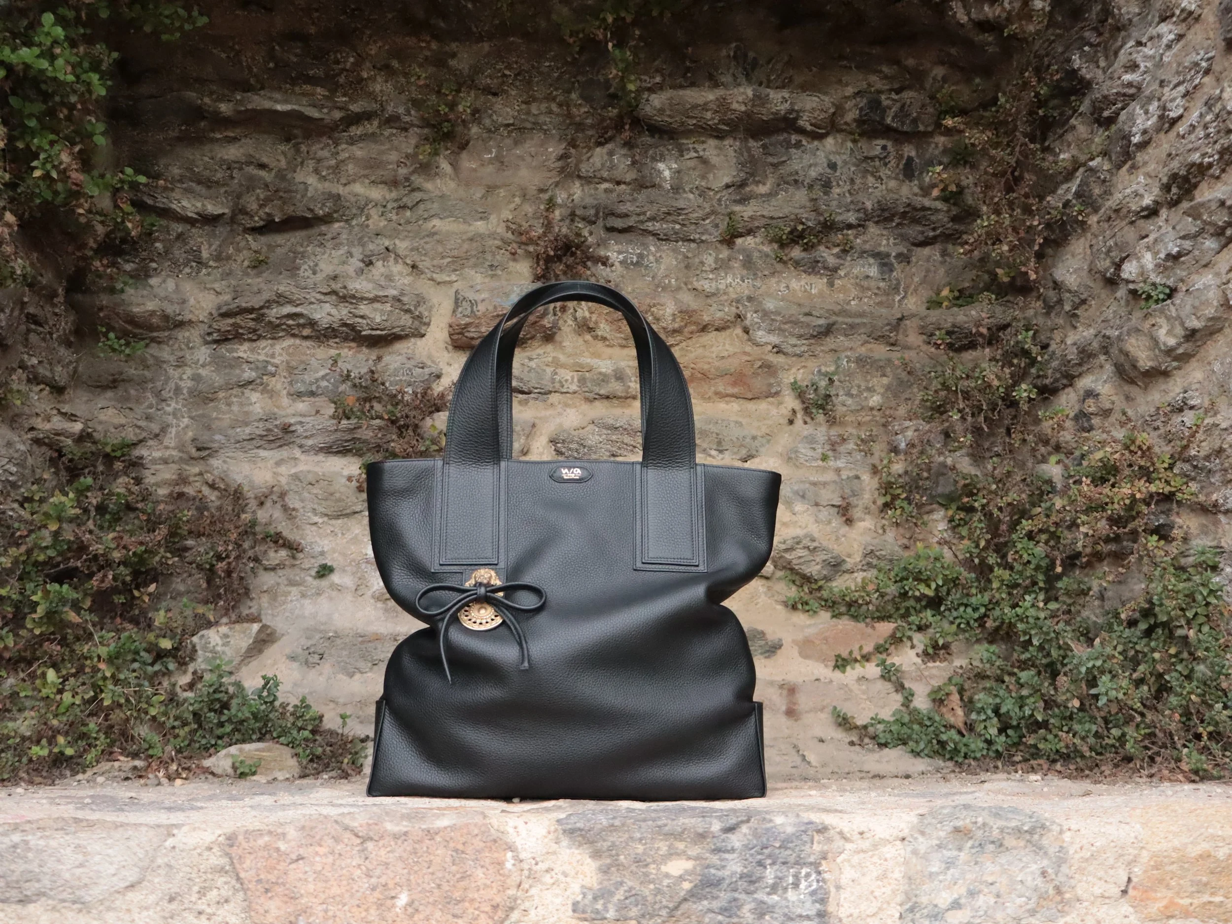 Sac à main noir en cuir posé sur un rebord en pierre devant un mur de pierre avec des plantes vertes