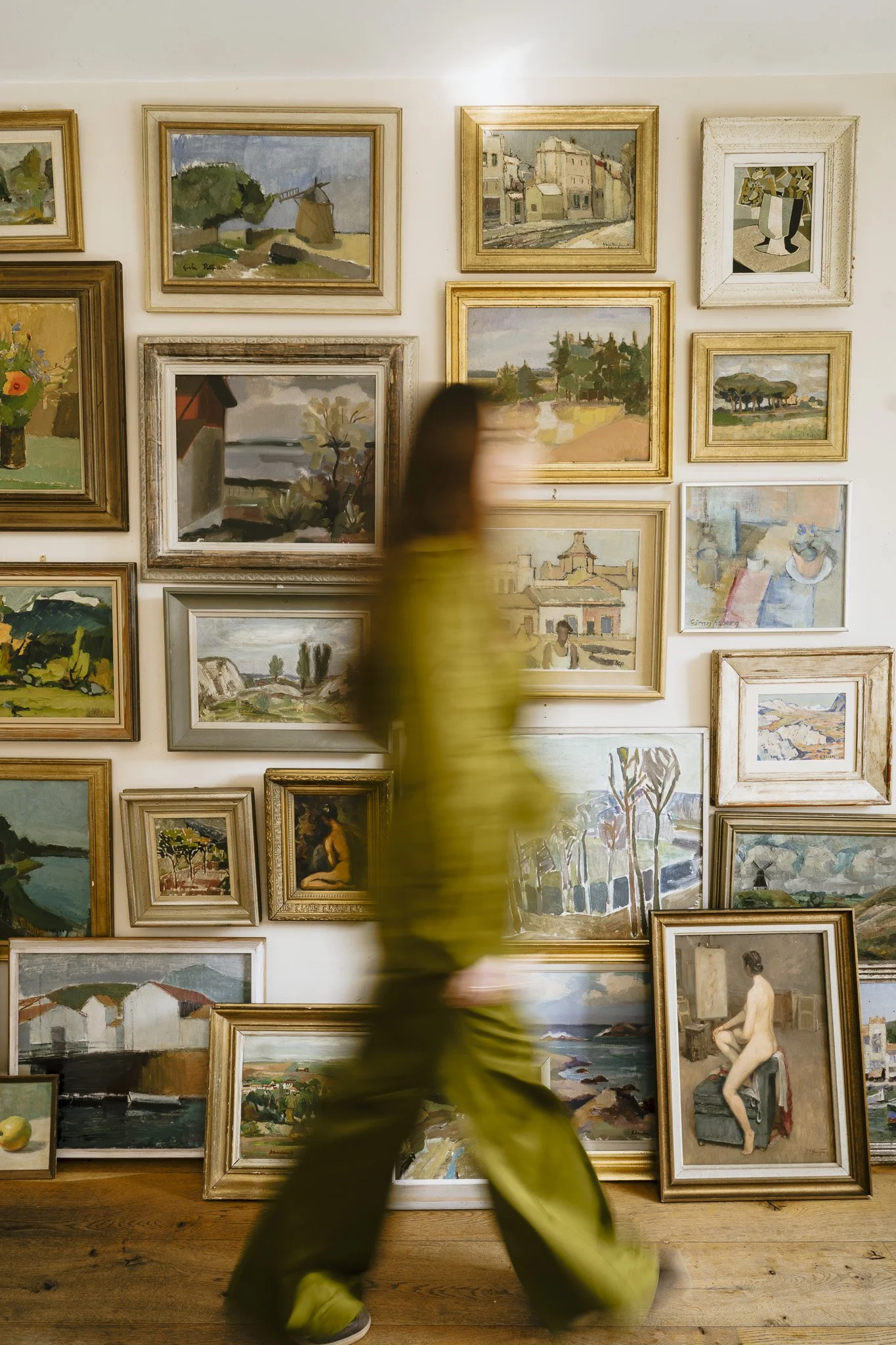 Blurry person walking past an art gallery wall filled with various landscape paintings and a portrait of a woman sitting on stairs.