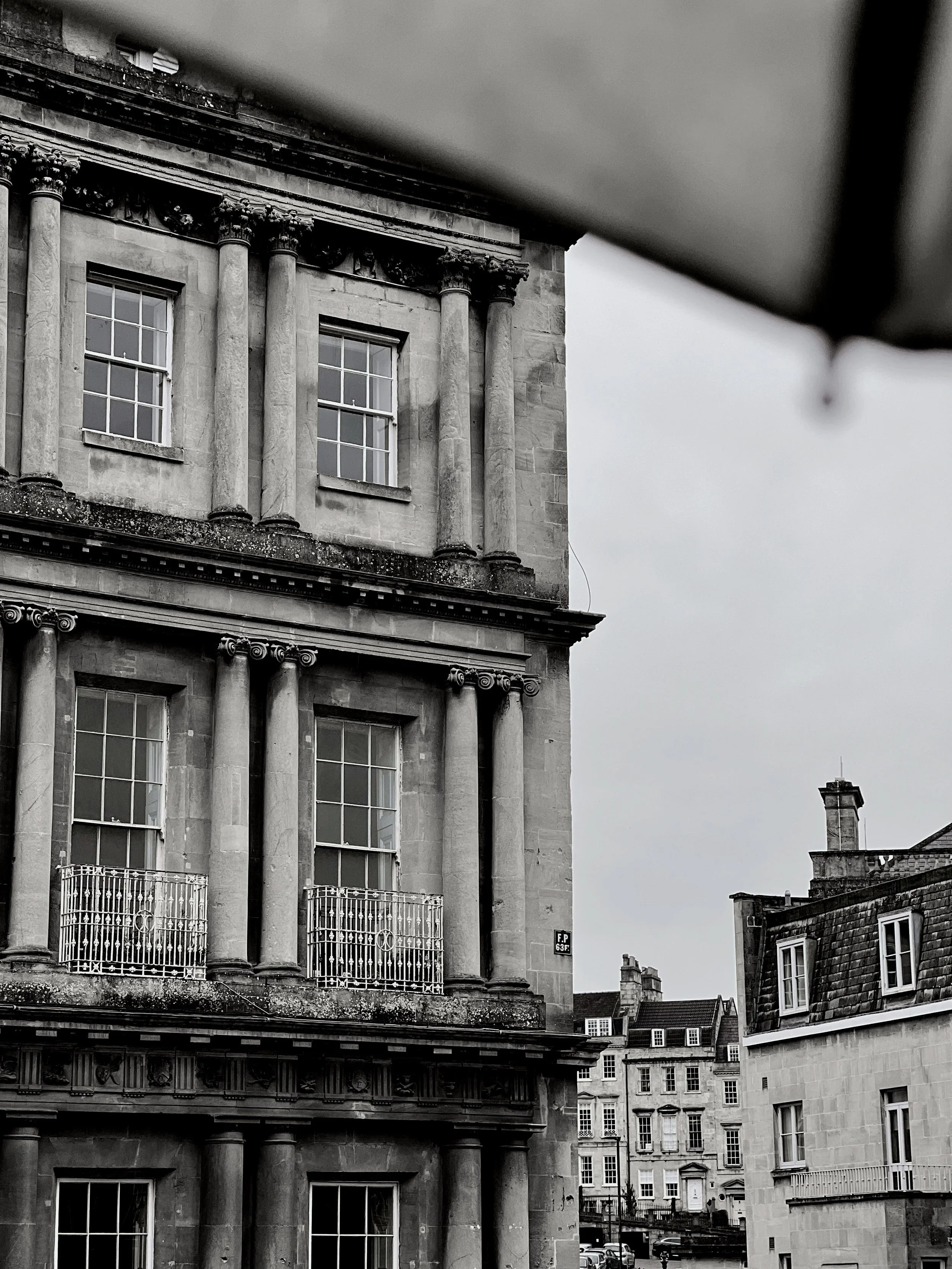 A black and white photo of historic European-style buildings with classical architectural elements such as columns, ornate friezes, and large windows, set in an urban area under an overcast sky.