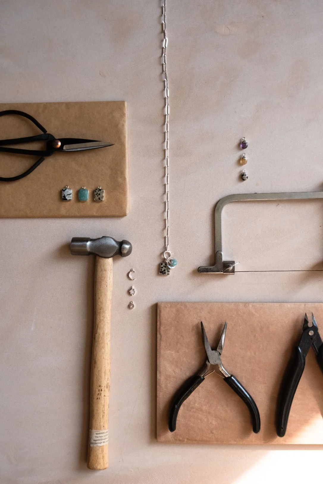 Jewelry making tools and supplies on a workspace, including pliers, a hammer, necklace chain, pendants, and beads.