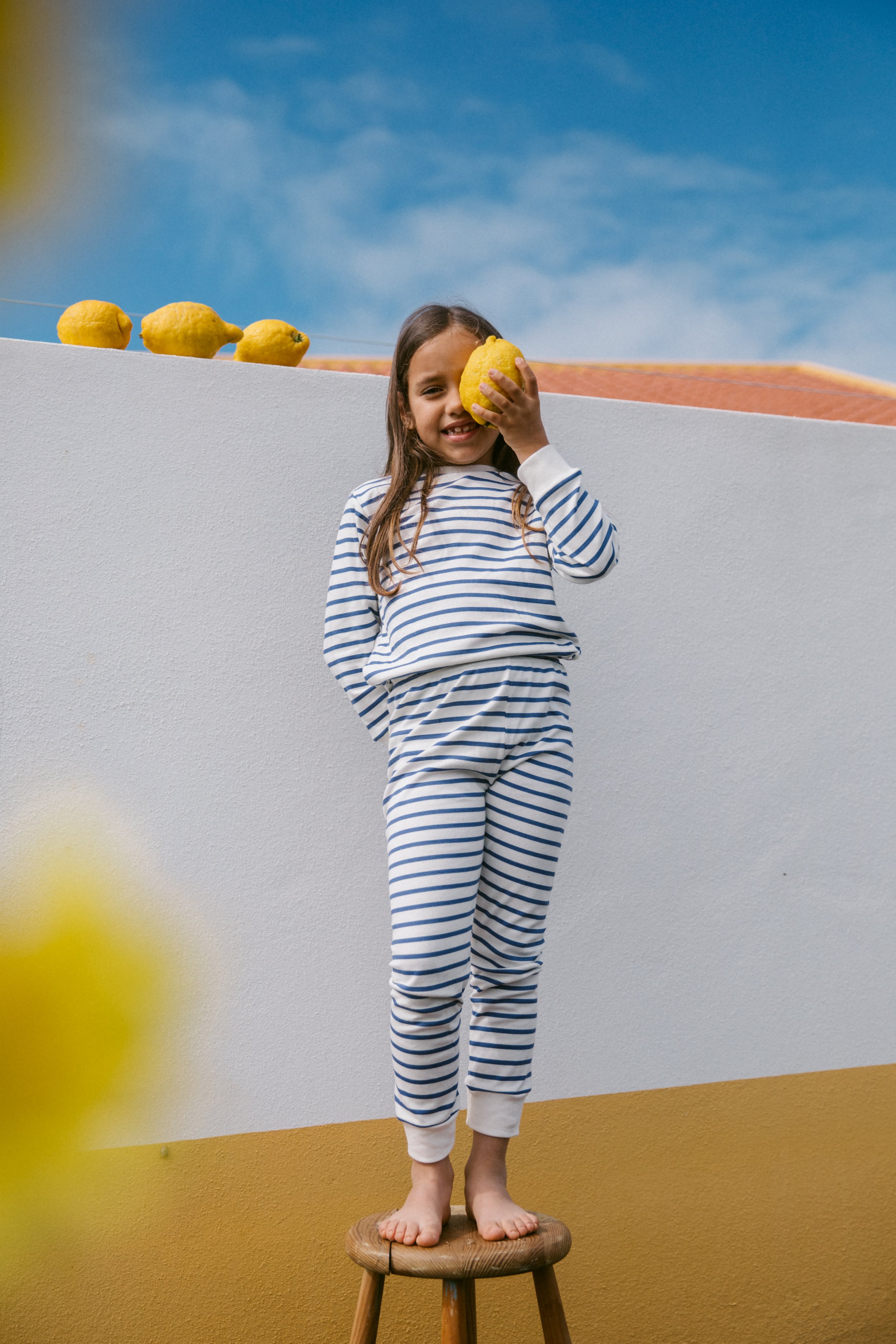 A young girl with long brown hair, wearing blue and white striped pajamas, standing barefoot on a wooden stool, holding a yellow lemon to her face, with a white and yellow wall behind her and a blue sky above. Several more lemons are lined up on a st