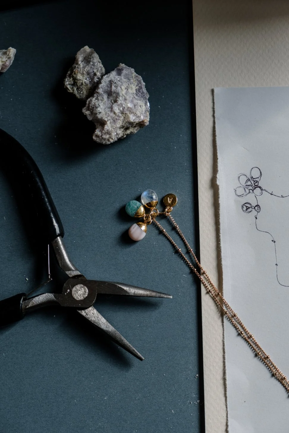 Jewelry-making tools and materials, including a pair of pliers, rocks, and a chain, arranged on a dark surface with a sketch on paper.