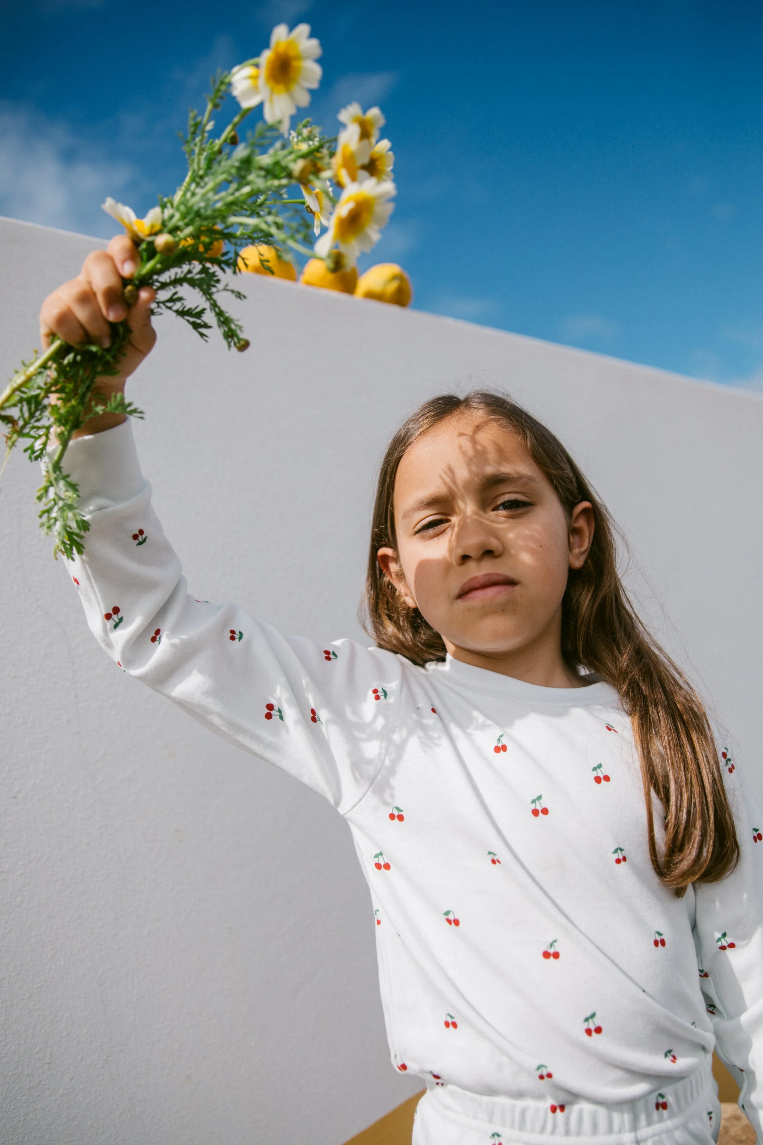 A young girl with long brown hair holding a bouquet of daisies and yellow fruits, standing outdoors against a modern white wall with a blue sky in the background.