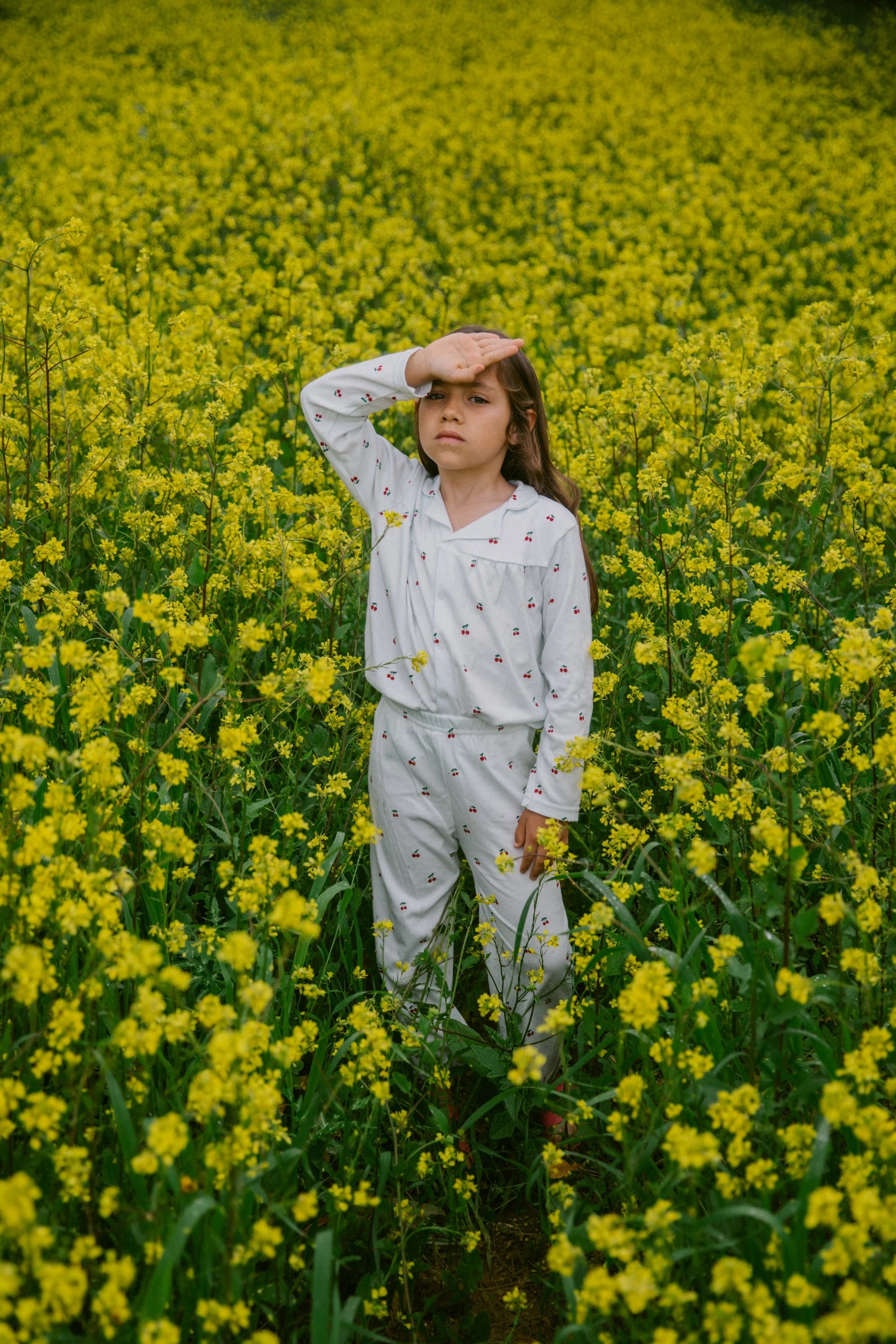 Young girl in pajamas standing in a yellow flowering field, shielding her eyes from the sun with her hand.