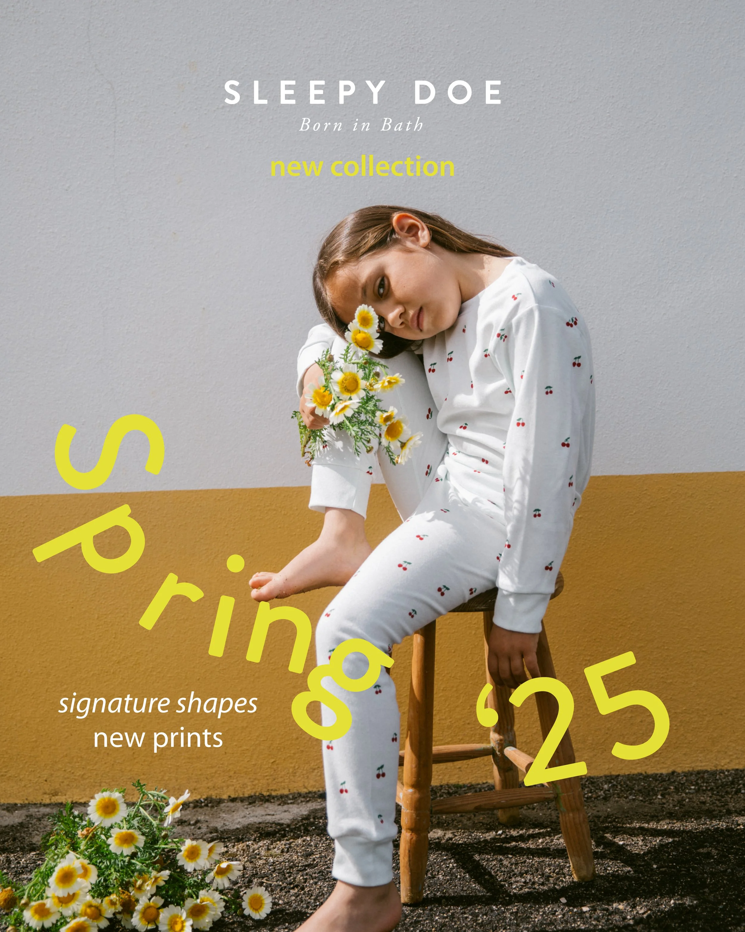 A woman sitting on a wooden chair outdoors against a two-tone wall, holding a bouquet of white daisies with yellow centers. She wears a white matching pajama set with a cherry print and has brown hair. There is yellow text overlaying the image that reads 'signature shapes new prints' and large pink and yellow partial text in the background.
