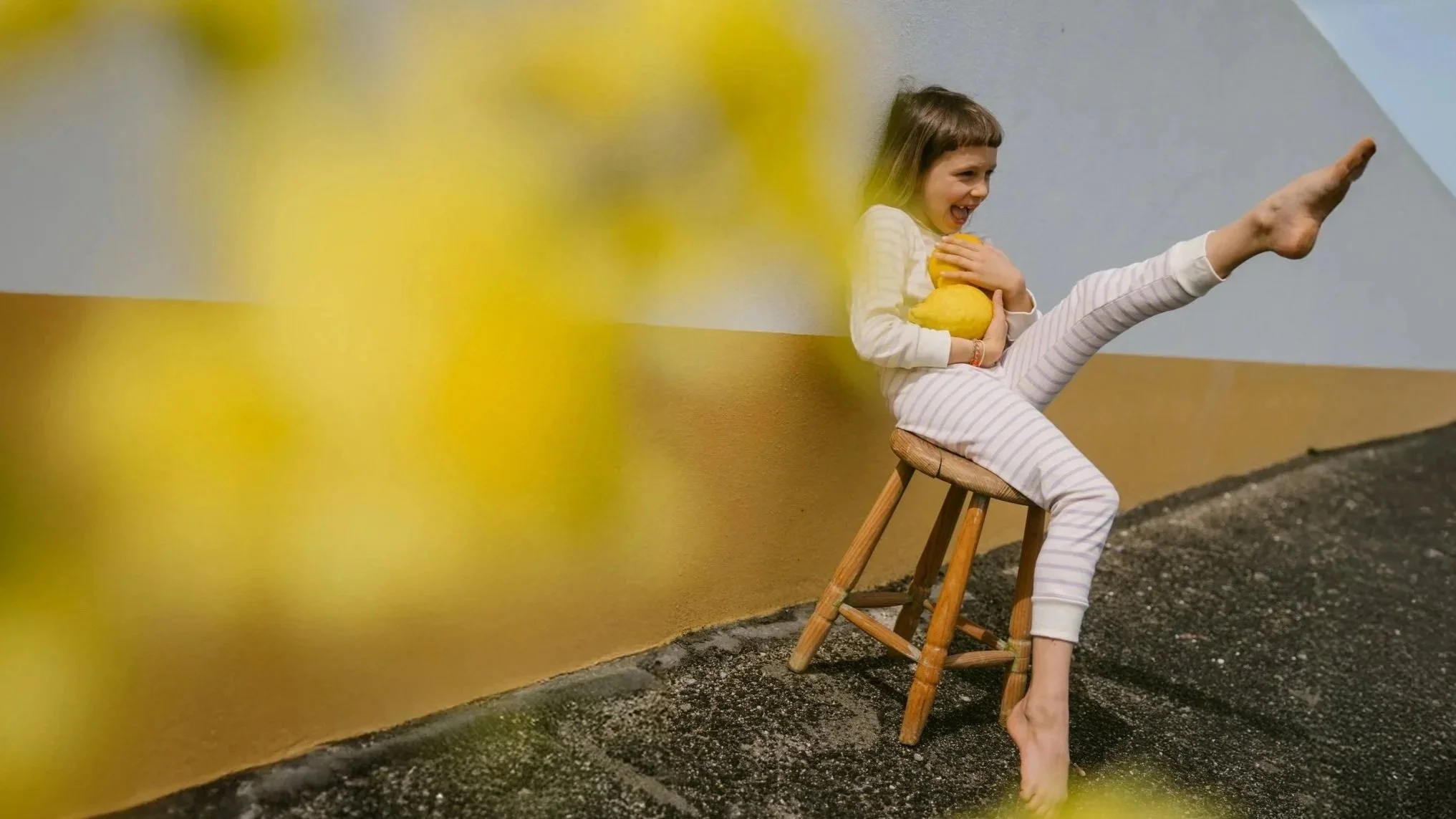 A young girl sitting on a wooden stool, holding a lemon, with one leg raised high and her foot pointing forward, smiling and laughing, against a two-toned colorful background.