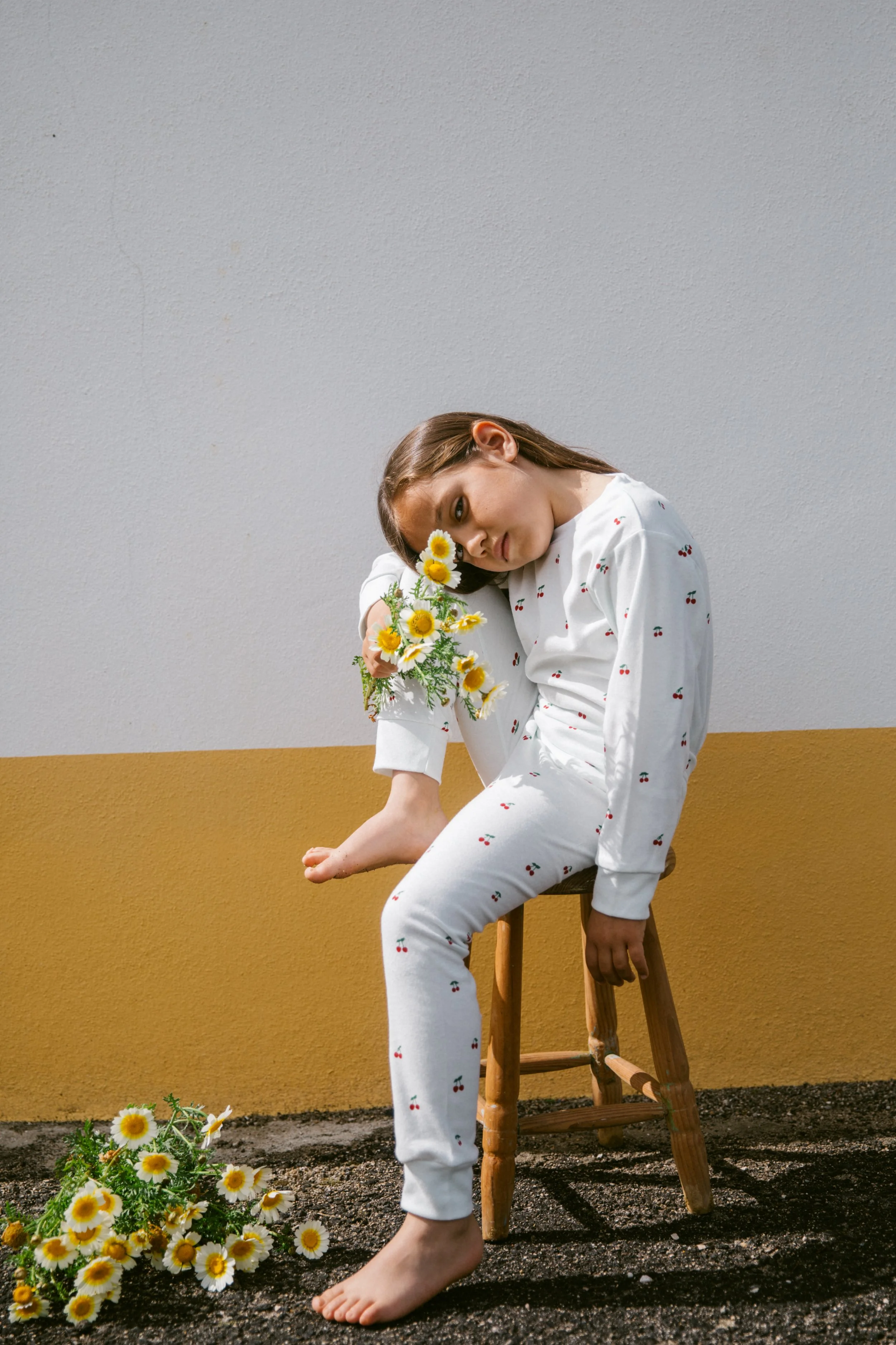 A young woman in white pajamas with small cherry patterns is seated on a wooden stool against a two-tone wall. She is leaning her head on a bouquet of yellow and white daisies, which she holds in her right hand. Her left foot is bare and resting on d