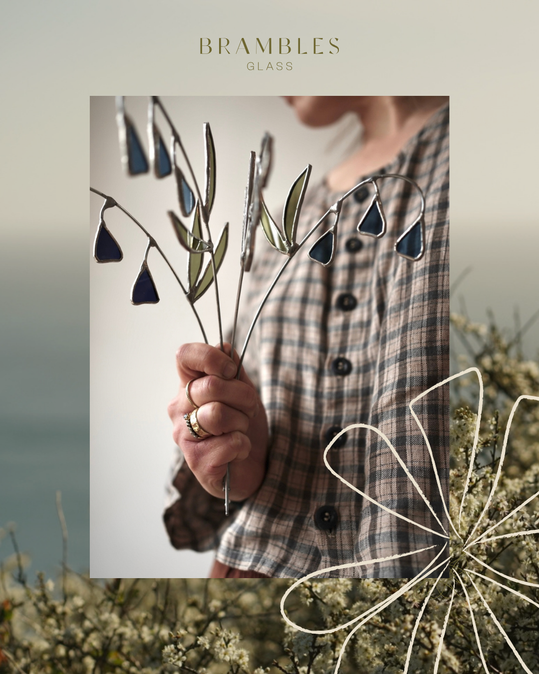 A person holding stained glass flower and leaf ornaments in front of a backdrop of white flowers and a blurred green background.