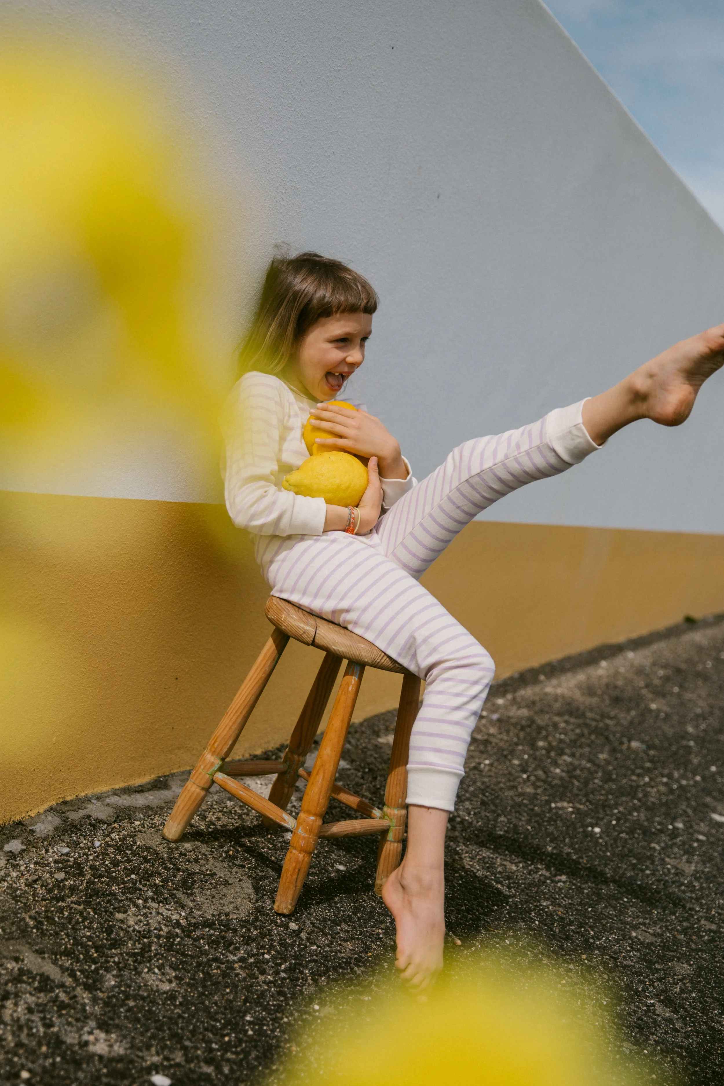 A girl with short brown hair wearing a striped pajama set and holding lemons is sitting on a wooden chair, raising one leg high and smiling, against a multi-colored wall with a blue sky visible at the top.