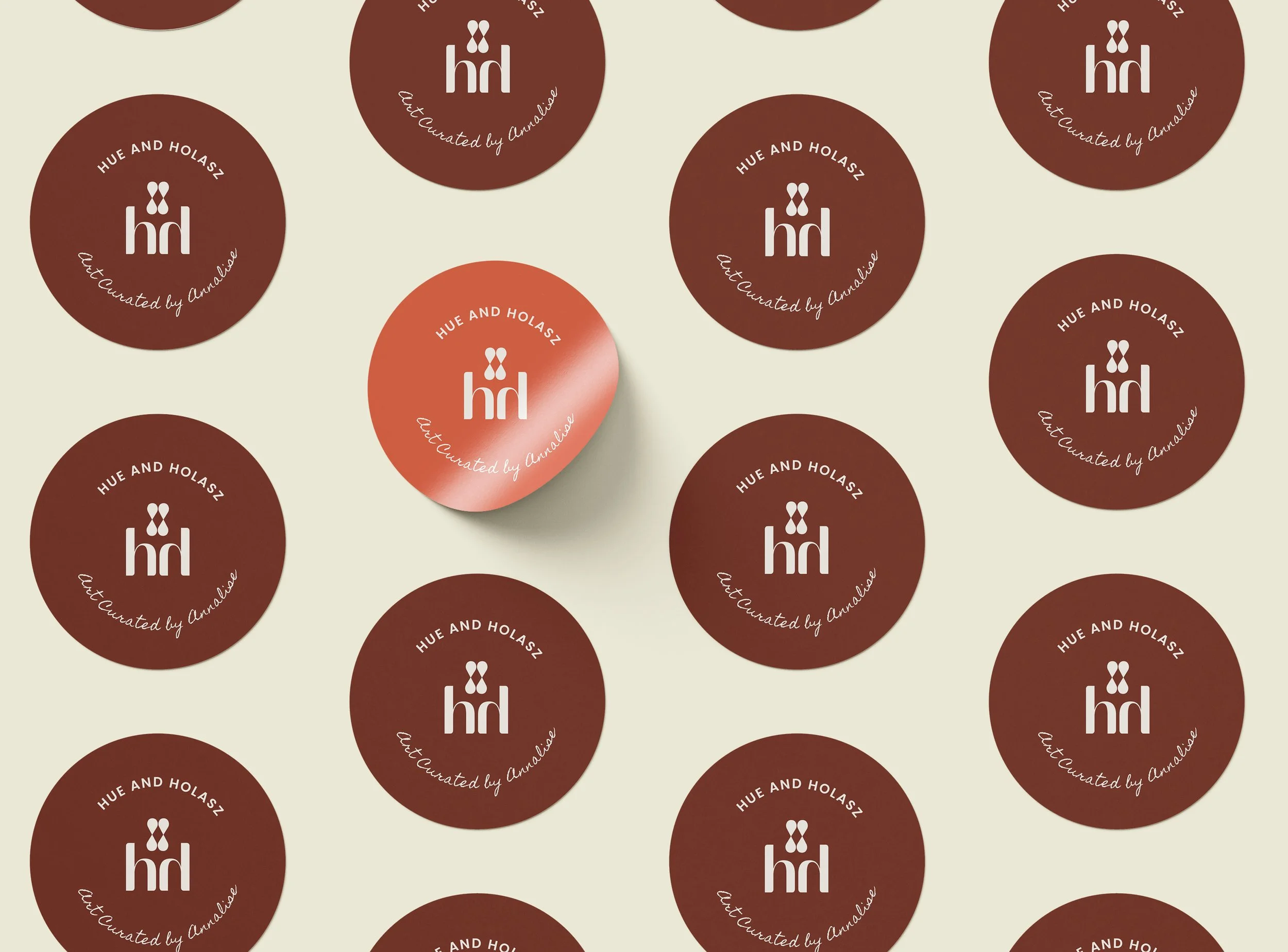 A pattern of round labels with a brown background, white text, and a logo. The labels have the words 'HUE AND HOLASZ' and 'art Curated by Amaleise' surrounding a white logo with the letters 'hd' and two small figures above.